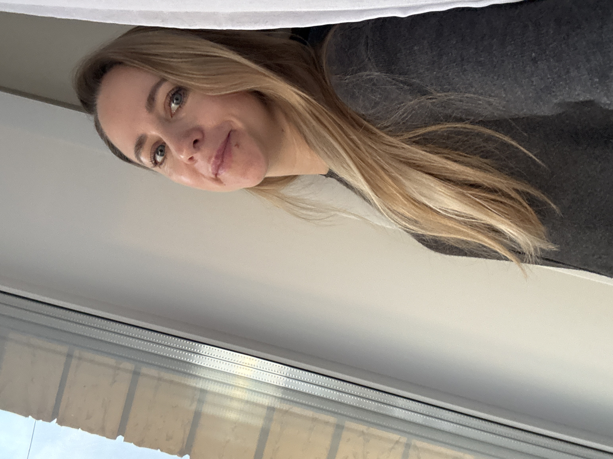 A portrait-style photograph captures a young woman positioned on the right side of the frame, looking towards the left. She has long, straight blonde hair, fair skin, and light blue eyes. Her expression is a subtle, playful smile, and her eyes appear curious or amused. A small, dark mole is visible just above her upper lip on the right side. She is wearing a dark gray top, partially visible.
A prominent white horizontal surface runs across the top of the image, obscuring the very top of her head. Below the woman, a light-colored wall or surface fills the mid-ground, transitioning into a window that occupies the bottom portion of the frame. The window features multiple horizontal divisions, and its glass reflects what appears to be part of an outdoor building or structure. The lighting is soft and even, suggesting an indoor setting illuminated by natural light, likely from the window. There is no visible text in the image. This scene is set in Chaikovsky, Russia.