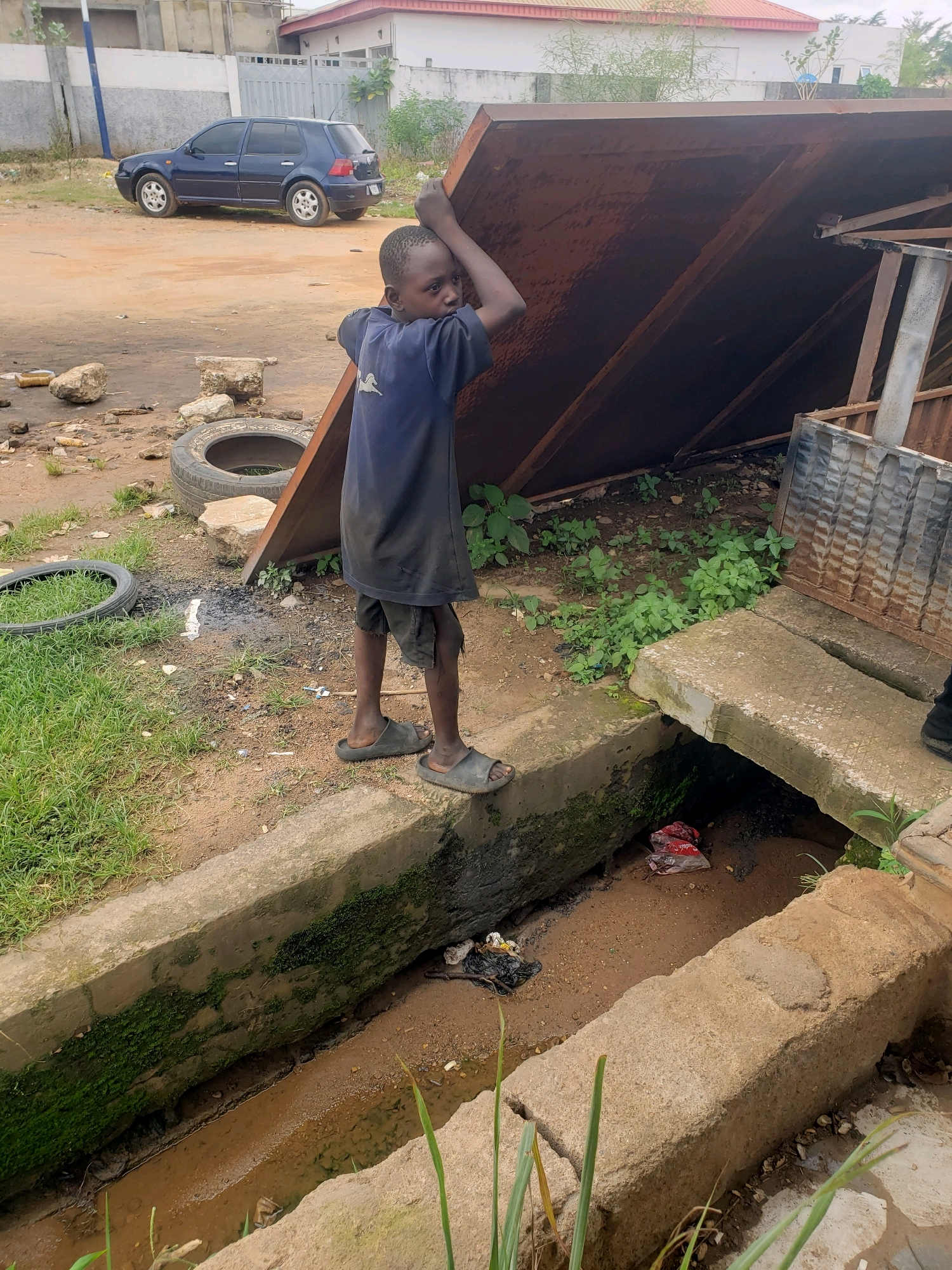 In Sabon Tasha, Nigeria, a young boy stands by a drainage ditch, his hands resting on a large, rusty metal sheet propped diagonally against a structure. He is wearing a dark blue t-shirt and dark shorts, and dark sandals. The ditch, made of concrete, is filled with muddy water and debris, including a red plastic bag with white lettering and some black trash. Green moss grows on the sides of the concrete structure. In the background, a dark blue Volkswagen Golf car is parked on a dirt road. Scattered around the area are discarded tires, rocks, and other assorted debris. Lush green grass grows in patches on the left side of the image.