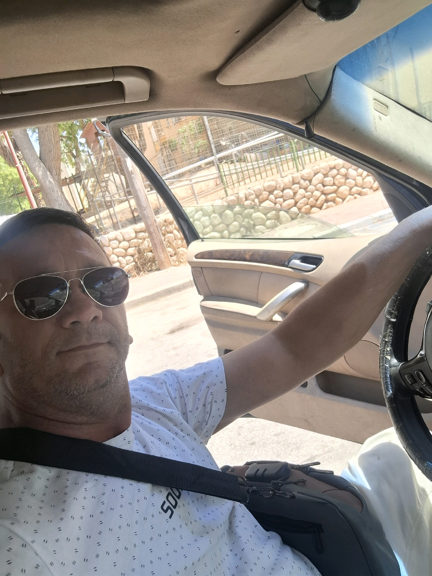 The image is a first-person perspective selfie taken from inside a car, likely in Dimona, Israel, given the arid, rocky landscape visible through the open car door and window. The driver, a man wearing sunglasses and a white patterned t-shirt, is positioned in the driver's seat, with his left arm resting on the steering wheel. His right arm is extended, possibly indicating he is holding the phone to take the selfie. The car's interior is visible, including the dashboard, door panel with a wooden trim, and the passenger seat. Outside, the scene depicts a sun-drenched environment with trees, stone walls, and a fence, consistent with a location in Israel. The man appears to be looking directly at the camera, with a neutral expression.