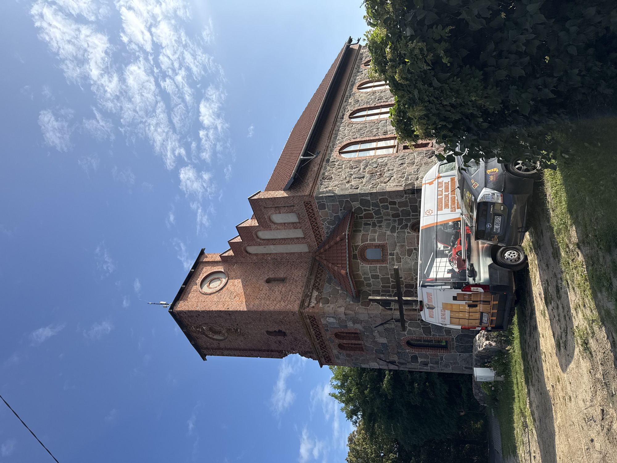 In Stare Brynki, Poland, a stone and brick building with a tiled roof and a prominent tower stands under a bright blue sky with scattered white clouds. Two vehicles are parked in front of the building on a dirt and grassy area. A white van with orange and black lettering and graphics of boxes and a red car is parked closest to the building. To the right of the van, a dark-colored sedan is partially visible, obscured by a lush green bush. The building exhibits architectural details such as arched windows and decorative brickwork. The overall scene suggests a daytime setting with clear weather.