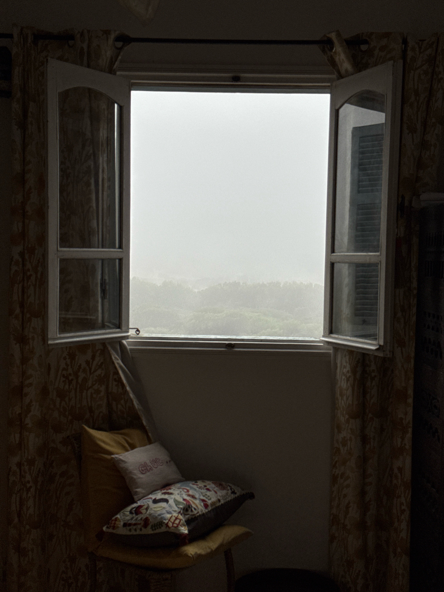 The image depicts a view from an open window in a room, looking out at a hazy landscape. The window itself is white-framed and divided into two panes by a horizontal bar. The left pane is fully open, revealing patterned curtains. The right pane is partially open, showing a glimpse of horizontal blinds. The window overlooks a dense, green landscape that fades into a misty background. In the foreground, inside the room, there are several pillows stacked on a cushioned seat. The pillows are of various colors and patterns, one of which has red lettering that appears to spell "CLUB..". The lighting is dim, suggesting it is either early morning or overcast. The scene is likely from a residential building in Essaouira, Morocco, given the architectural style and the presence of patterned textiles common in the region. There are no people or animals visible in the image.