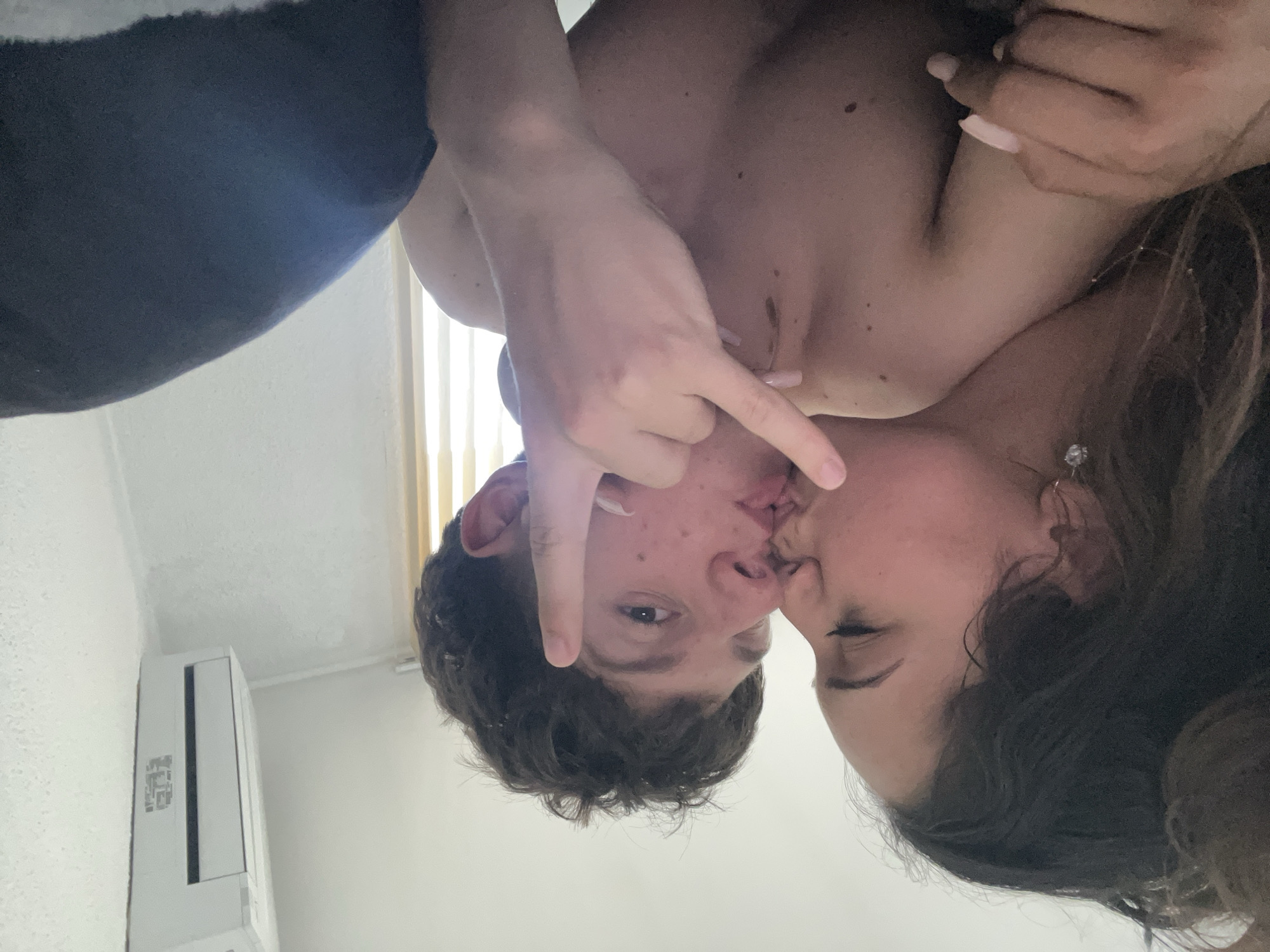 A close-up, horizontal interior photograph captures two individuals engaged in an intimate interaction in what appears to be a bedroom in Zlatograd, Bulgaria. On the left, a female, with dark, curly hair partially visible, is depicted in profile, kissing a male on the lips. She wears a small, sparkling stud earring in her left ear and a delicate necklace. Her left hand, adorned with light pink, elongated nails, rests on the male's right shoulder. The male, positioned centrally, faces slightly towards the viewer with his eyes open. He has short, dark, curly hair and appears shirtless, revealing visible moles/blemishes on his chest. His right hand is raised to the right side of his head, forming a gesture with his index and pinky fingers extended upwards, also displaying light pink, elongated nails. In the upper right background, a white wall-mounted air conditioning unit is visible. Vertical window blinds are partially seen on the right side of the image, behind the male, indicating a light source from that direction. The lower right foreground shows a dark blue or black textile, possibly a blanket or pillow. The walls are light-colored with minor textural variations.