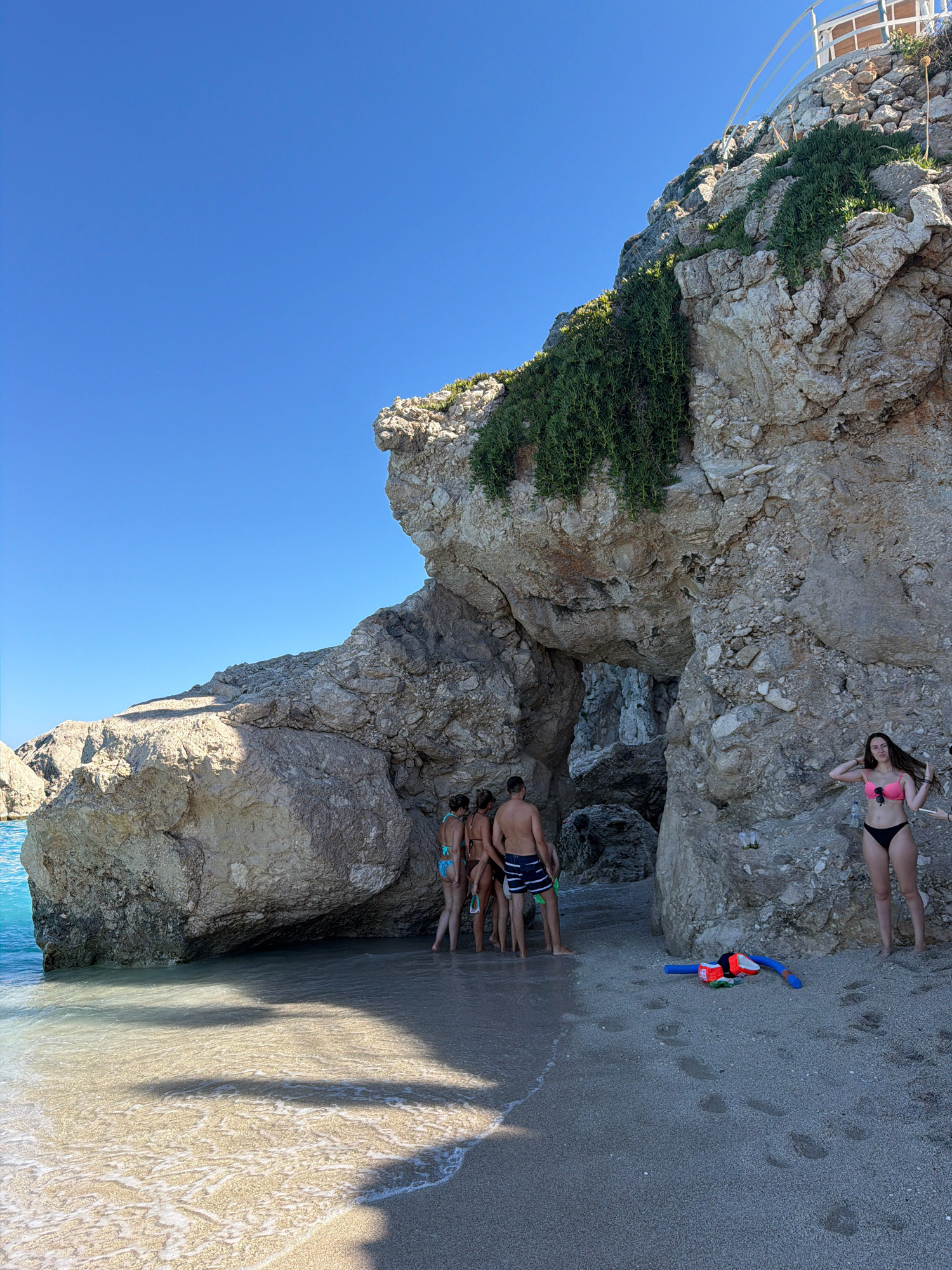 In the Municipal Unit of Lefkada, Greece, a group of six people are on a sandy beach next to a large rocky cliff formation. The cliff has an opening that forms a natural archway or cave. The sky is clear blue.
On the beach to the left, four individuals, three women and one man, are standing near the water. The women are wearing bikinis, and the man is wearing swim trunks. They appear to be looking towards the rocky opening.
To the right, a woman in a pink bikini and black bottoms is standing on the sand, looking towards the camera. She has her hands near her hair. In the foreground, a snorkel and mask set, with a blue tube and orange and black accents, is lying on the sand. A small life vest or flotation device is also present. Further to the right, another person's leg is visible, suggesting another individual is present but mostly out of frame.
The water is clear and turquoise, with gentle waves lapping at the shore. Sunlight casts shadows on the sand and water. The rocky cliff is textured with visible strata and some green vegetation growing on its upper parts.