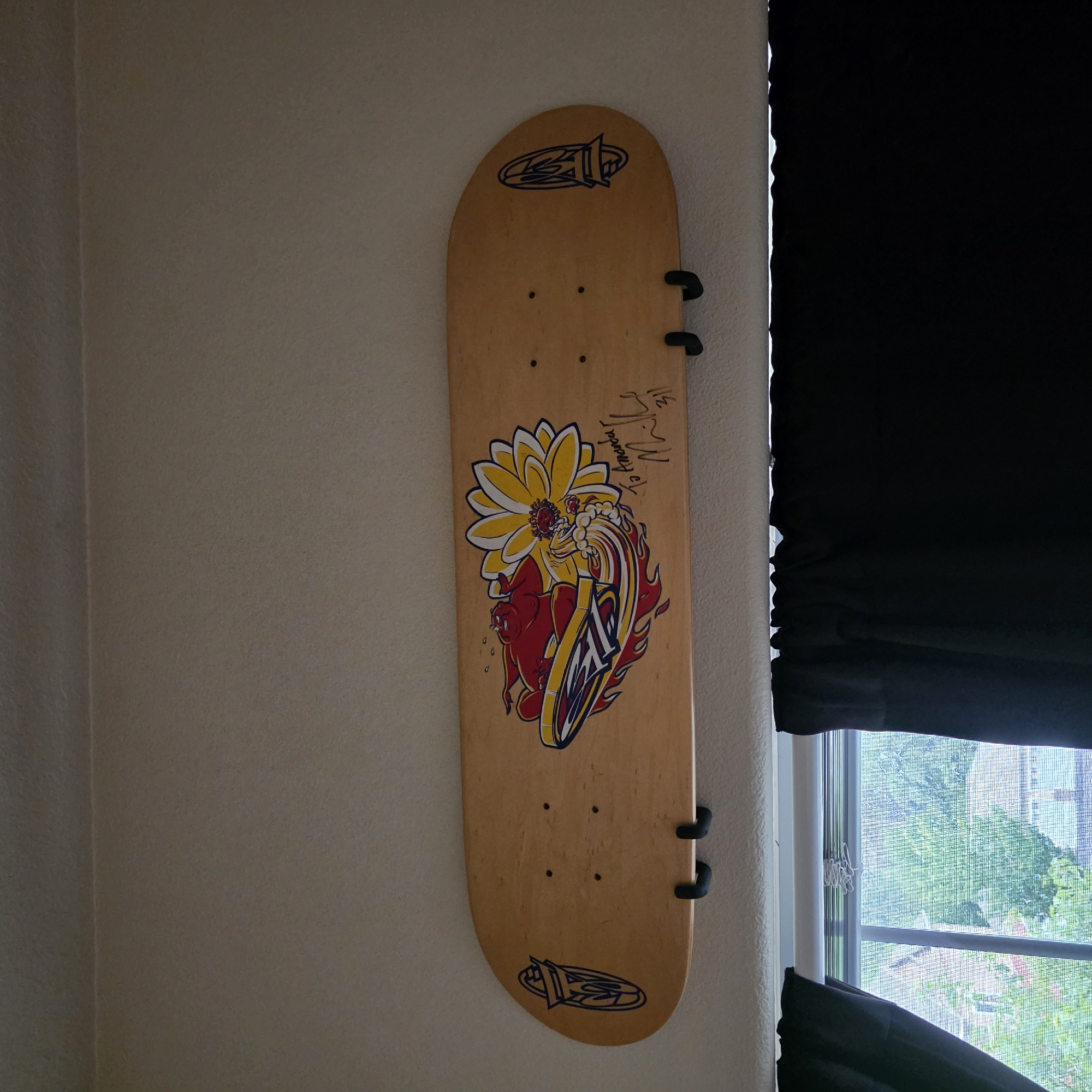 This media file shows a wooden skateboard mounted vertically on a textured, off-white wall. The skateboard has a graphic design featuring a yellow flower with red and white accents, and a red figure in a surfing pose with flames and a yellow surfboard with a blue logo. The skateboard also has two dark clips attached to it, presumably for mounting. There is a signature on the right side of the skateboard, above the graphic, with the text "Jeffrey Wet" and some other illegible writing. The top and bottom of the skateboard also feature a blue logo in an oval shape.
To the right of the skateboard, a black curtain is partially drawn, revealing a window with a view of trees and buildings outside. The sunlight streaming through the window suggests it is daytime. The setting appears to be an indoor space, possibly a bedroom or living area, in Beaverton, United States of America. The overall mood is casual and displays a piece of skateboarding memorabilia.