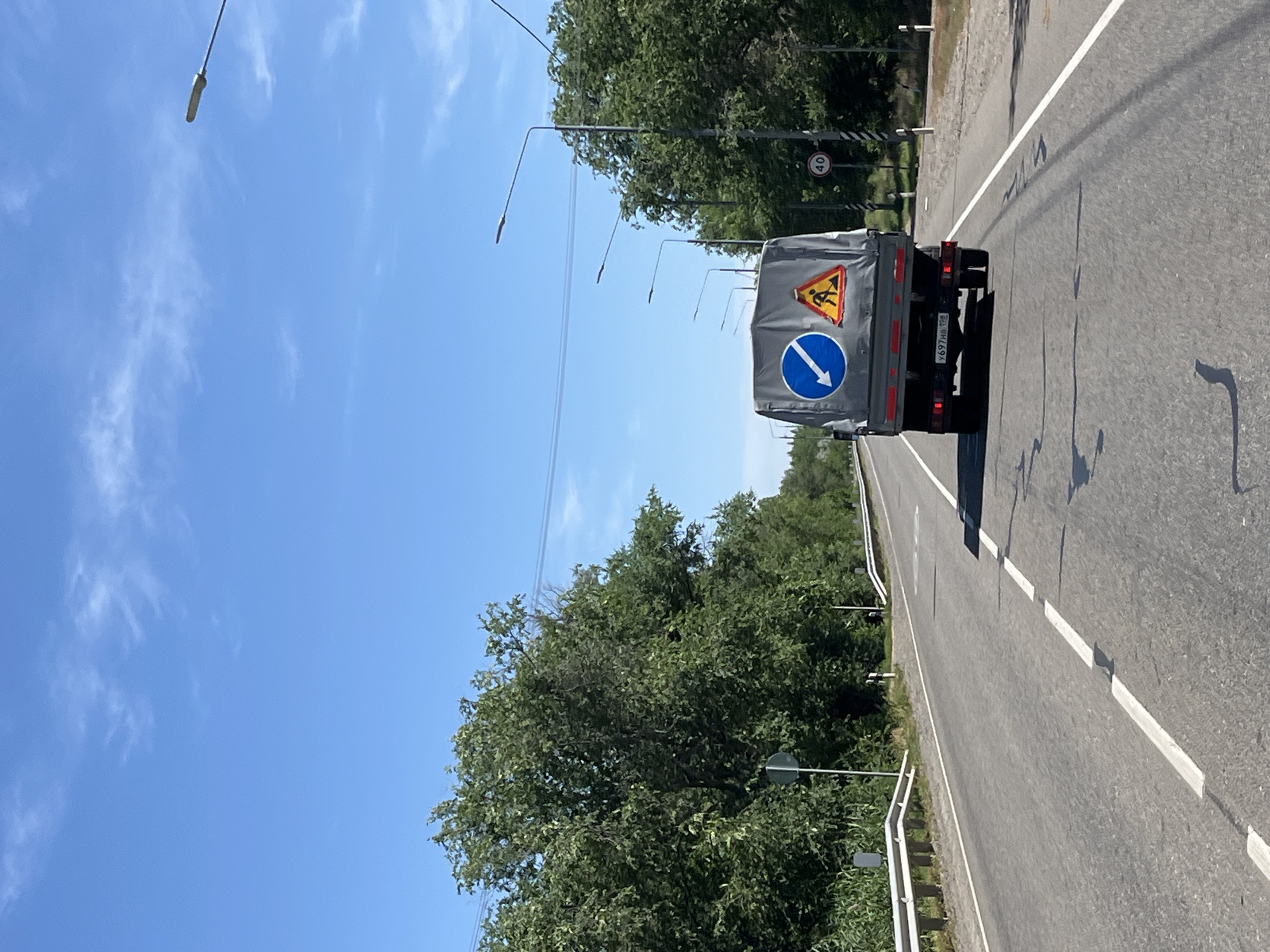 The image shows a truck driving on a two-lane asphalt road. The truck is covered with a grey tarp and has two traffic signs on its back: a blue circular sign with a white arrow pointing down and to the left, and a yellow triangular sign depicting a person working. The truck's license plate is visible and reads "У697НА 199". On the right side of the road, there is a circular speed limit sign displaying "40". The road is lined with trees and vegetation on both sides. The sky is blue with a few wispy clouds. Several streetlights are visible along the road. The scene appears to be a rural or suburban area, possibly on the outskirts of Alqaza, Russia, given the presence of the license plate format.