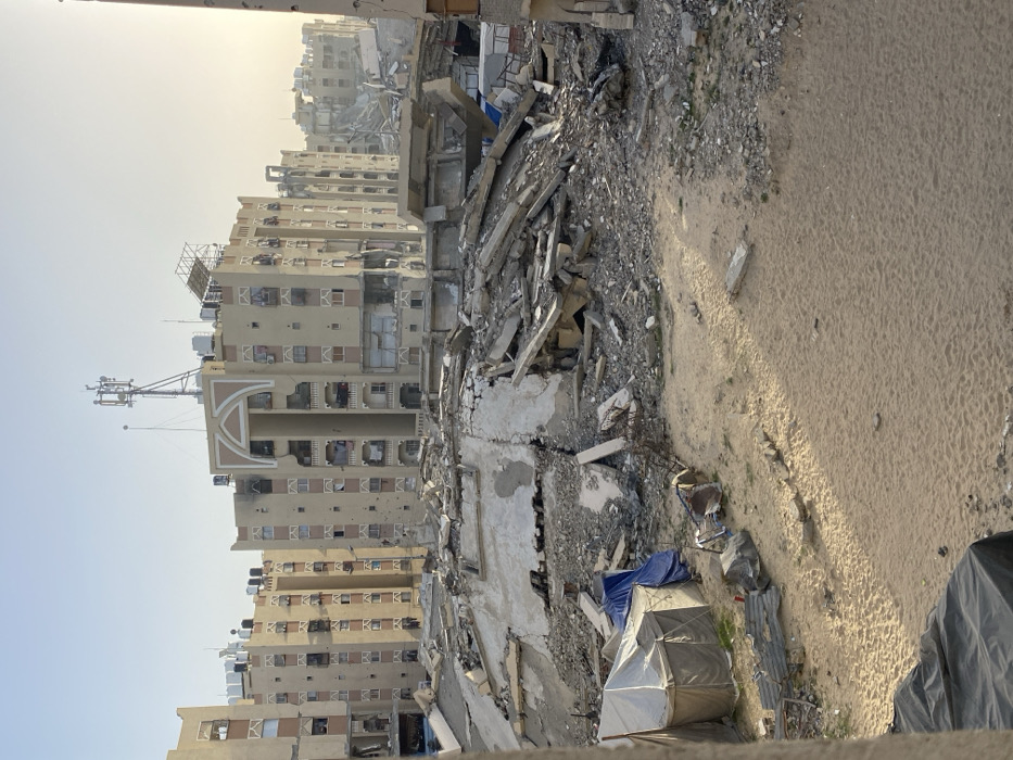 The image depicts a scene of extensive urban destruction, characterized by collapsed buildings and widespread debris, set under a clear, bright sky. The perspective is slightly elevated, looking down and across the landscape.
In the immediate foreground, a significant amount of rubble and shattered concrete dominates the left side, indicating a completely destroyed structure. This debris consists of broken concrete slabs, twisted rebar, and smaller fragments of building materials. To the right of this rubble, the ground transitions to a sandy or dusty expanse, which shows subtle linear patterns, possibly tire tracks or footpaths.
Towards the bottom center and right, nestled amidst the debris and on the edge of the sandy area, are several makeshift structures. A blue tarp is visible, covering an unseen object. Adjacent to it are two tent-like shelters made of light-colored fabric or tarp material, suggesting temporary dwellings. One is larger, and the other is smaller, placed closer to the foreground. A dark grey tarp is partially visible along the far bottom right edge. Scattered pieces of debris, including what appears to be a metal frame or object, are near these shelters.
Beyond the immediate foreground, a flattened area with remnants of a foundation or floor slab is visible, also covered in scattered rubble. In the midground and background, a series of multi-story residential buildings stand, showing various degrees of damage. These buildings are predominantly light-colored (beige or off-white) and appear to be apartment blocks. Many windows are missing or shattered, and dark marks, consistent with impact damage or holes, are visible on their facades. Some balconies are damaged or partially collapsed.
On the roof of one of the more prominent buildings in the mid-left, a tall communications tower or antenna mast with multiple dishes and aerials is clearly visible. Other rooftops also feature water tanks or utility structures. Further into the background, more buildings can be seen, some appearing damaged, receding into a light haze near the horizon.
The sky is bright and clear, with a noticeable glare or haze in the upper left portion of the image. Shadows cast on the sandy ground suggest that the sun is relatively low, indicating either morning or late afternoon. The overall impression is one of devastation and displacement in an urban environment. No discernible text is visible in the image.