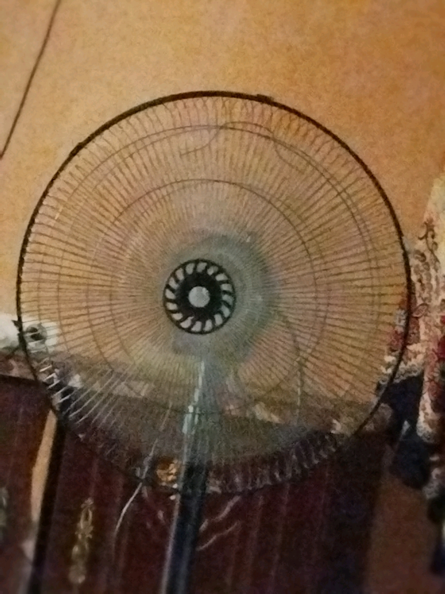 The image shows a close-up, slightly blurred view of an oscillating fan. The fan is positioned in the center of the frame, facing forward. Its grille is made of thin, closely spaced metal wires, forming a circular pattern with a darker, circular hub in the center. The hub has a decorative pattern of dark and light segments. The fan's stand is visible below the grille, appearing to be made of dark metal. The background is out of focus and consists of a textured, light brown wall. To the right of the fan, a portion of a patterned fabric, possibly a curtain or tapestry, with floral motifs in red, white, and dark blue is visible. The overall lighting is dim, casting a warm tone over the scene. The location is identified as Tarakkiyot, Uzbekistan.