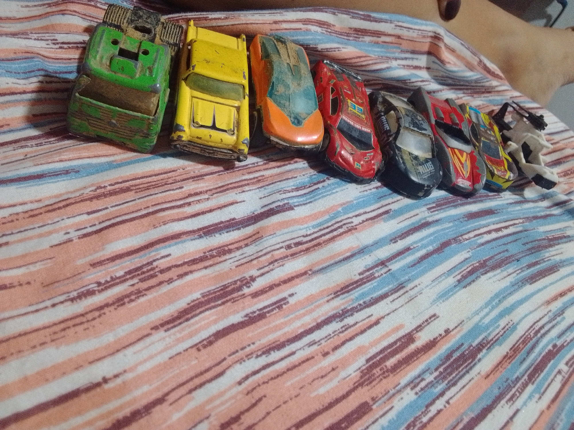 An overhead, medium shot shows a collection of toy cars arranged diagonally on a striped fabric, likely a bedsheet. The fabric features a pattern of thin, diagonal lines in shades of peach, white, maroon, and light blue. To the right, a person's bare legs and feet are visible, resting on the fabric. The toy cars are positioned from the top left to the bottom right of the frame. The car at the far left is a green, blocky vehicle with a visible dirt accumulation on its surfaces. It is followed by a yellow, rectangular car, then an orange and blue-colored car with a sleek design, and a red racing car with various decals. Further to the right, a black car with a silver stripe, another red car with yellow accents, a yellow car with red and black detailing, and a white and black toy motorcycle are visible. All the toy vehicles appear worn and dusty, suggesting they are old and have been played with. The scene appears to be indoors, on a bed.