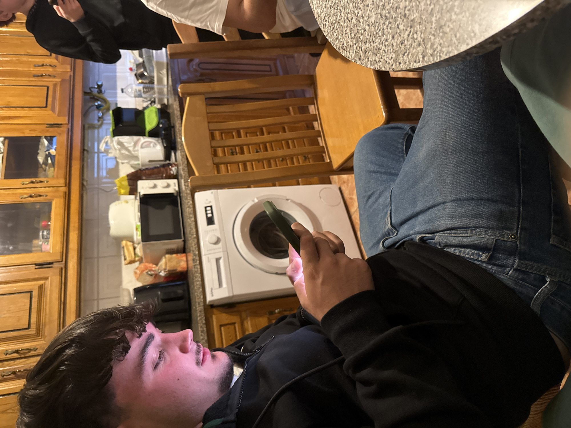A medium shot depicts a young man with dark hair and a mustache, wearing a black hoodie, looking at a smartphone in his hand. He is seated at a wooden chair at a table with a speckled surface. Adjacent to him is another person wearing jeans. In the background, there are wooden cabinets, a washing machine, and other kitchen appliances. The location is a kitchen interior, potentially in Bragança, Portugal, though that can't be definitively determined from the image. There are other people at the table as well.