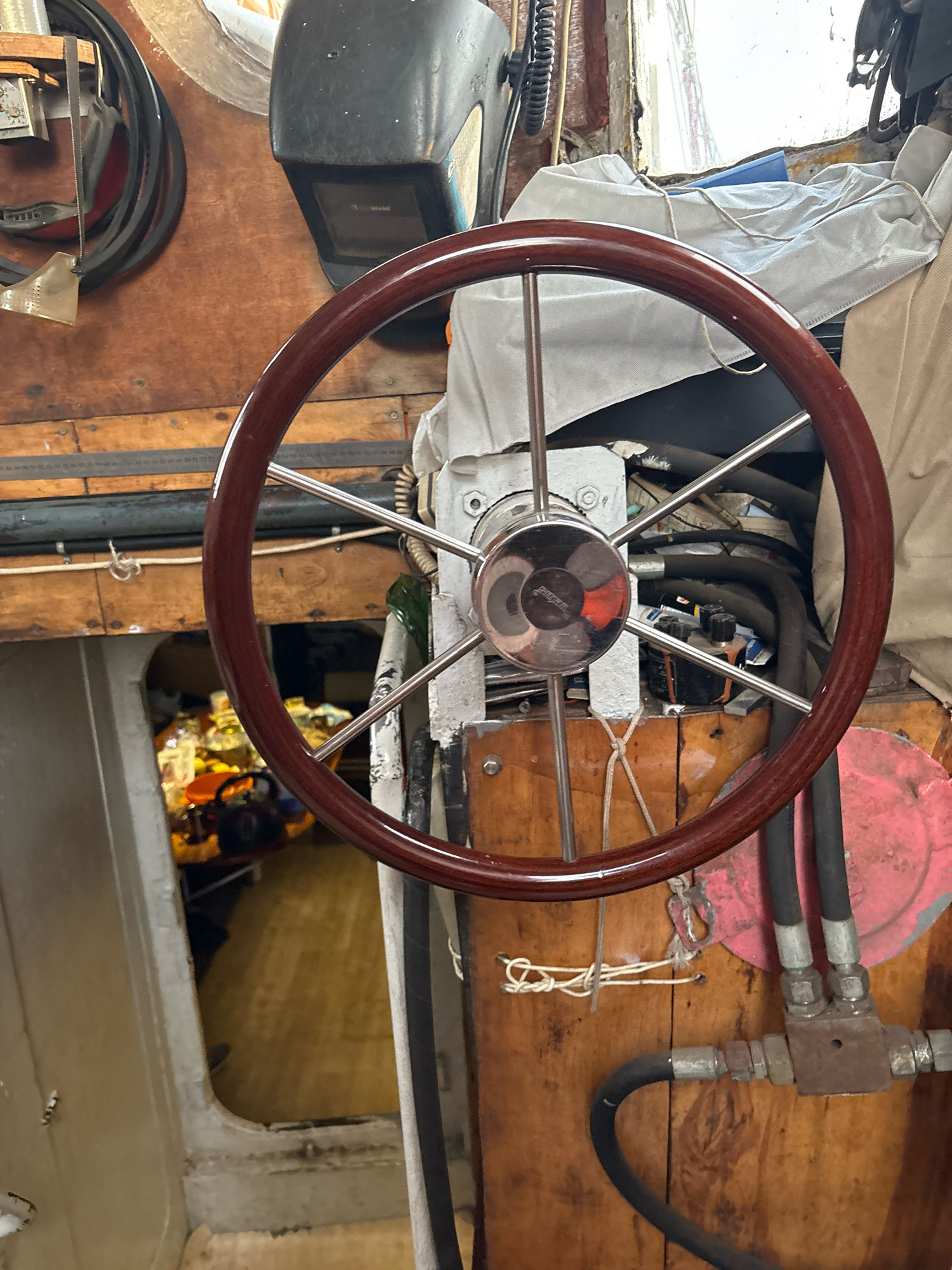 The image depicts the interior of a boat, specifically the helm area. A polished wooden steering wheel with metal spokes is centrally located. Behind the wheel, a complex system of black hoses and metal fittings is visible, suggesting hydraulic or mechanical equipment. The walls and surfaces appear to be made of wood, showing signs of age and wear. In the background, through an opening, a dimly lit area with various items, including what appear to be bottles and containers, can be seen. The overall environment appears utilitarian and possibly part of a working vessel. There are no people visible in the image. The scene is consistent with an indoor setting on a boat, possibly in a marina or docked.