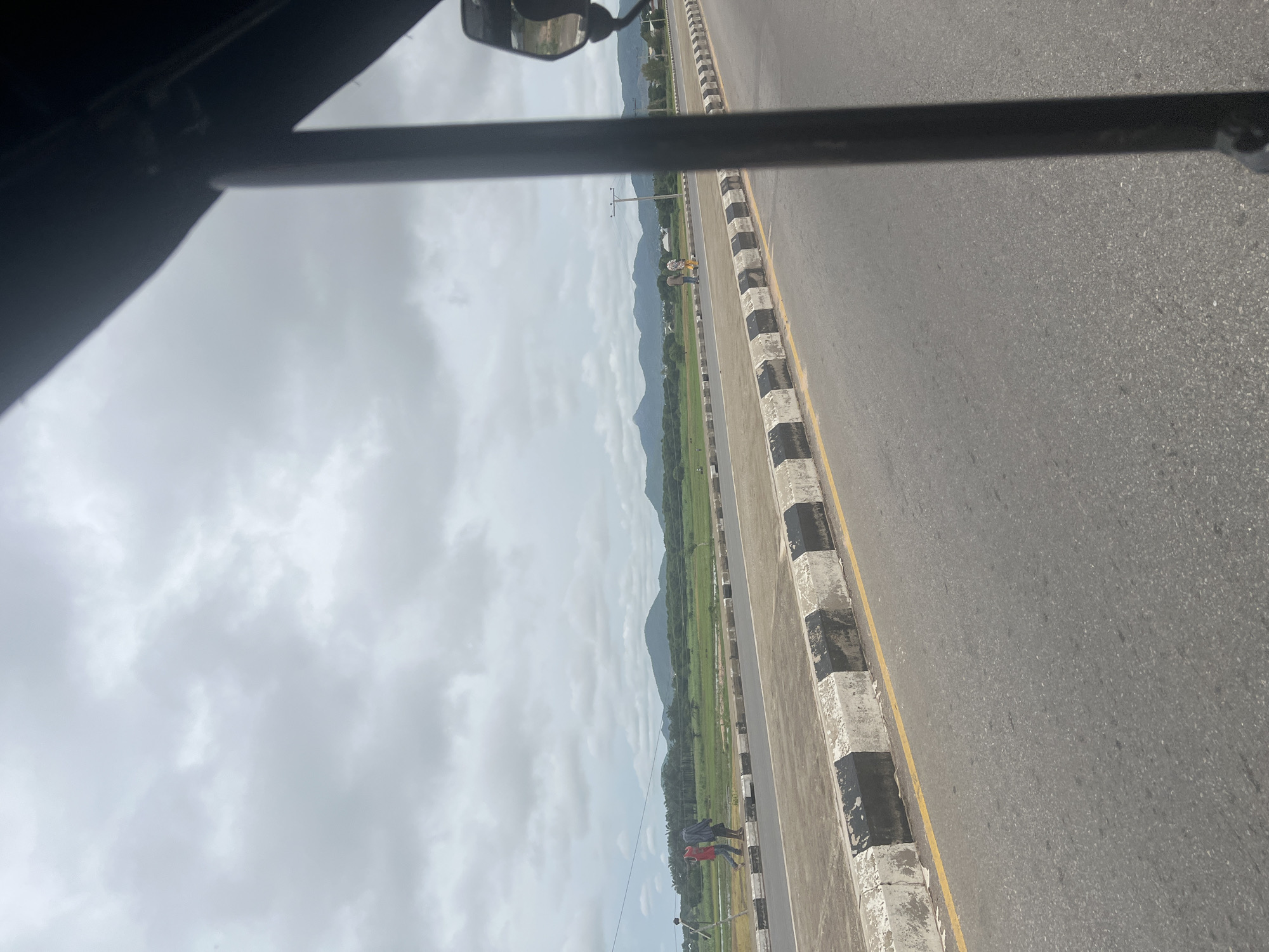 The image captures a view from inside a vehicle in Jalingo, Nigeria, looking out onto a paved road with a distinct black and white striped curb. Beyond the road, a grassy field stretches towards a range of green, undulating hills under a cloudy sky. Two individuals are visible walking on the pedestrian path beside the road in the mid-ground, and further in the distance, a few more figures can be seen. Power lines and poles are present along the roadside, suggesting an inhabited area. The overall scene depicts a semi-urban or rural environment on an overcast day.
