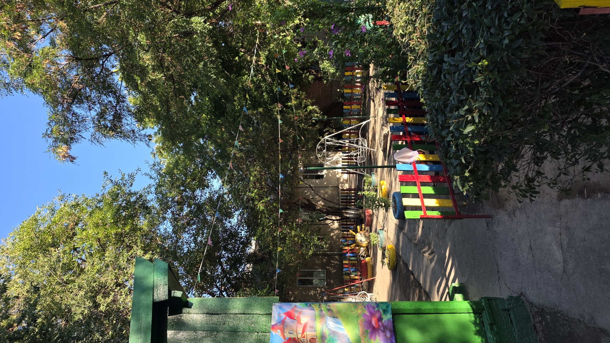 The image depicts an outdoor playground area, likely part of a kindergarten or similar facility, located in the city of Tokmok, Kyrgyzstan. The scene is bathed in bright sunlight, casting distinct shadows on a paved pathway. Lush green trees with dense foliage surround the playground, with one particularly large tree dominating the central area. The sky is a clear, vibrant blue.
Dominating the foreground on the left is a portion of a vibrant, green painted structure, possibly a building or a fence, adorned with a colorful mural depicting a fairytale castle with a waterfall and flowers. A low, brightly painted fence, featuring alternating red, yellow, blue, and green vertical slats, encloses parts of the playground. Beyond this fence, several playground elements are visible. A white metal swing set stands centrally, with a single swing attached. Other playground features include a tire transformed into a planter and painted to resemble a bee, and several other tire planters holding small green plants. In the background, a single-story building with several windows is visible, partially obscured by trees and fencing. Colorful triangular flags are strung between trees and structures, adding a festive atmosphere. Dense green bushes and foliage occupy the right side of the image, with some flowering vines visible. The overall impression is one of a well-maintained, cheerful outdoor space for children.