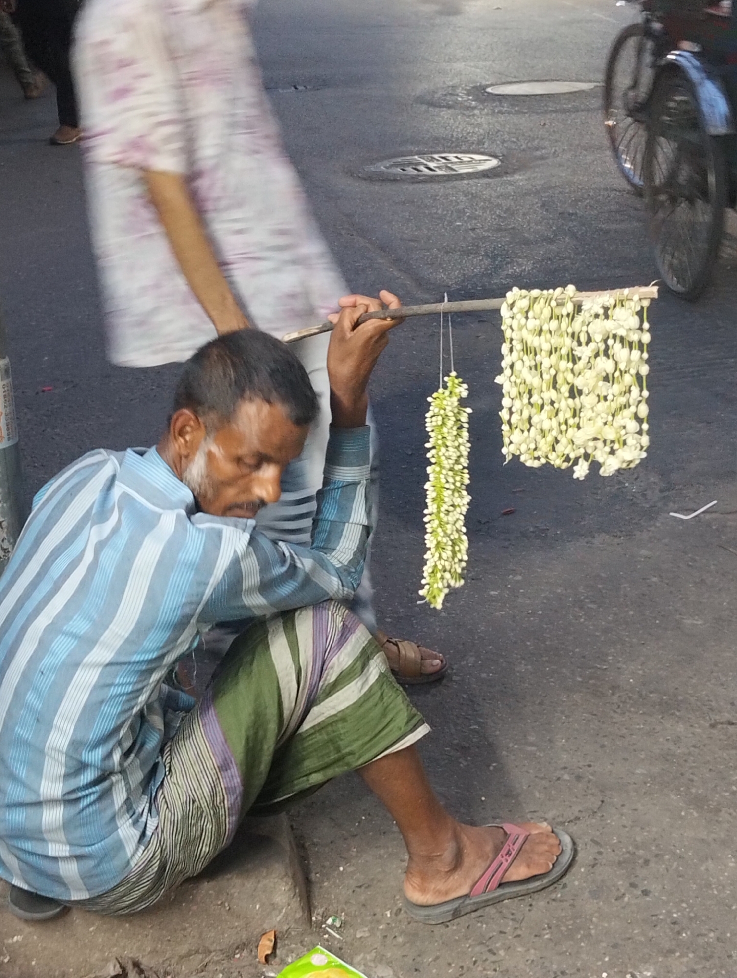 A street scene captures a man seated on the ground, holding a wooden stick from which strands of jasmine flowers are hung for sale. The man is positioned in the foreground, wearing a light blue and white striped long-sleeved shirt, green and purple striped lungi-style trousers, and mismatched sandals (one brown, one grey with a pink strap). He has dark hair with visible grey, and a beard with white patches on his cheeks, and he looks downwards, obscuring his facial expression.
From the wooden stick he holds, two different styles of jasmine garlands are displayed: a longer, denser strand of mostly closed white buds, and a shorter, wider garland of open white flowers.
The setting appears to be an urban street or sidewalk, with dark asphalt visible in the background featuring circular manhole covers. A blurred person in a light-colored garment with a faint pattern is seen moving in the background, along with a blurred bicycle wheel and fender on the right, suggesting passing traffic. Another blurry person's legs are partially visible on the far left. The lighting is soft, possibly indicating late afternoon or early evening.
On a metal pole or stand to the left of the man, some faint, illegible text is visible, including what appears to be a phone number: "01892-38995". A green and white packet lies on the ground near the man's left foot, and small pieces of debris are scattered on the rough ground surface. The overall image depicts a moment of everyday life, likely a street vendor at work.