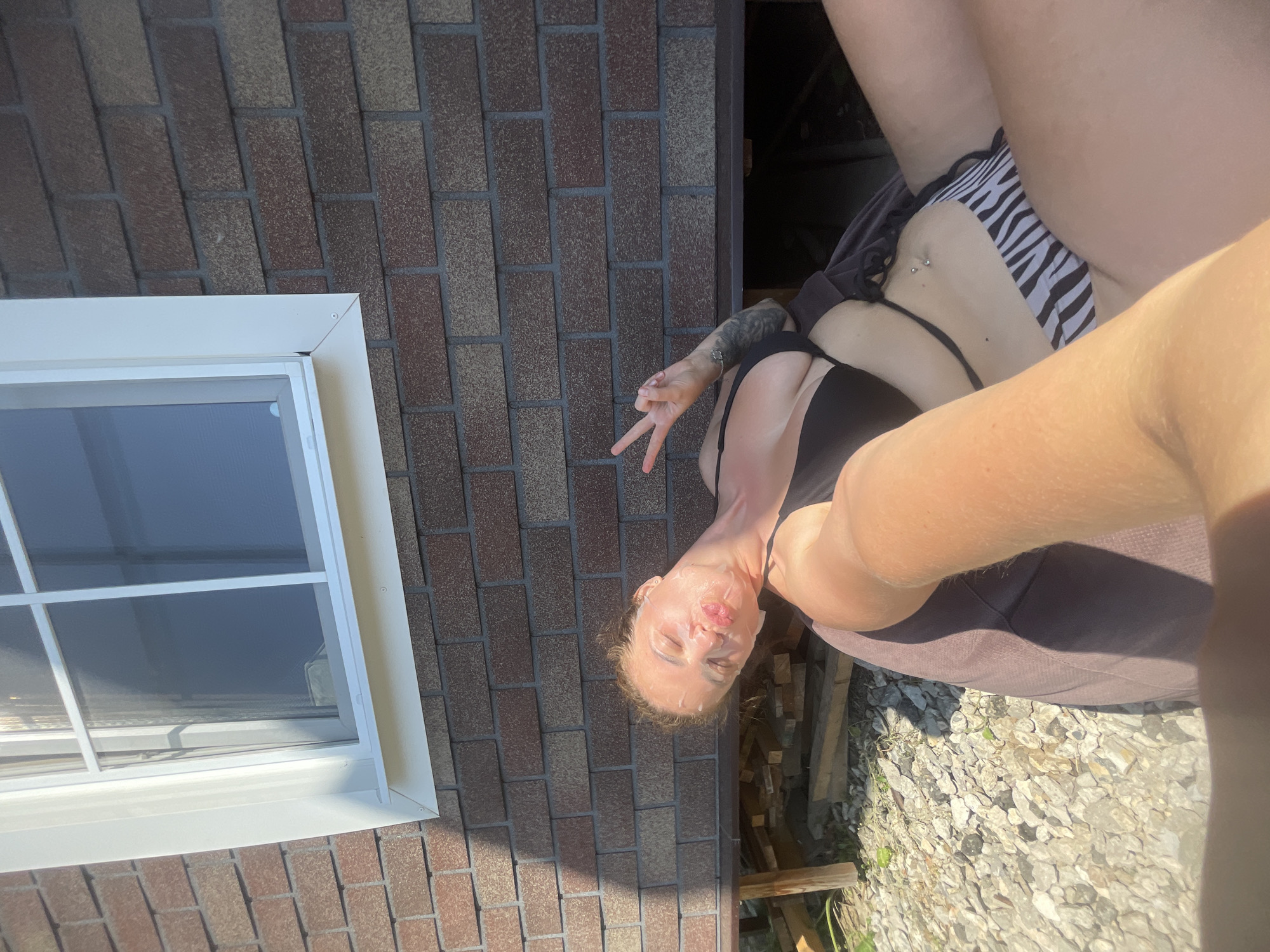A woman with a face mask is lying outside near a brick building. She is wearing a black bikini top and zebra-striped bikini bottoms. The building has a window with white trim. The ground is covered in gravel and there are some wooden planks visible. The woman is holding up two fingers in a peace sign. The lighting suggests it is daytime and sunny. The location is not discernible from the image.