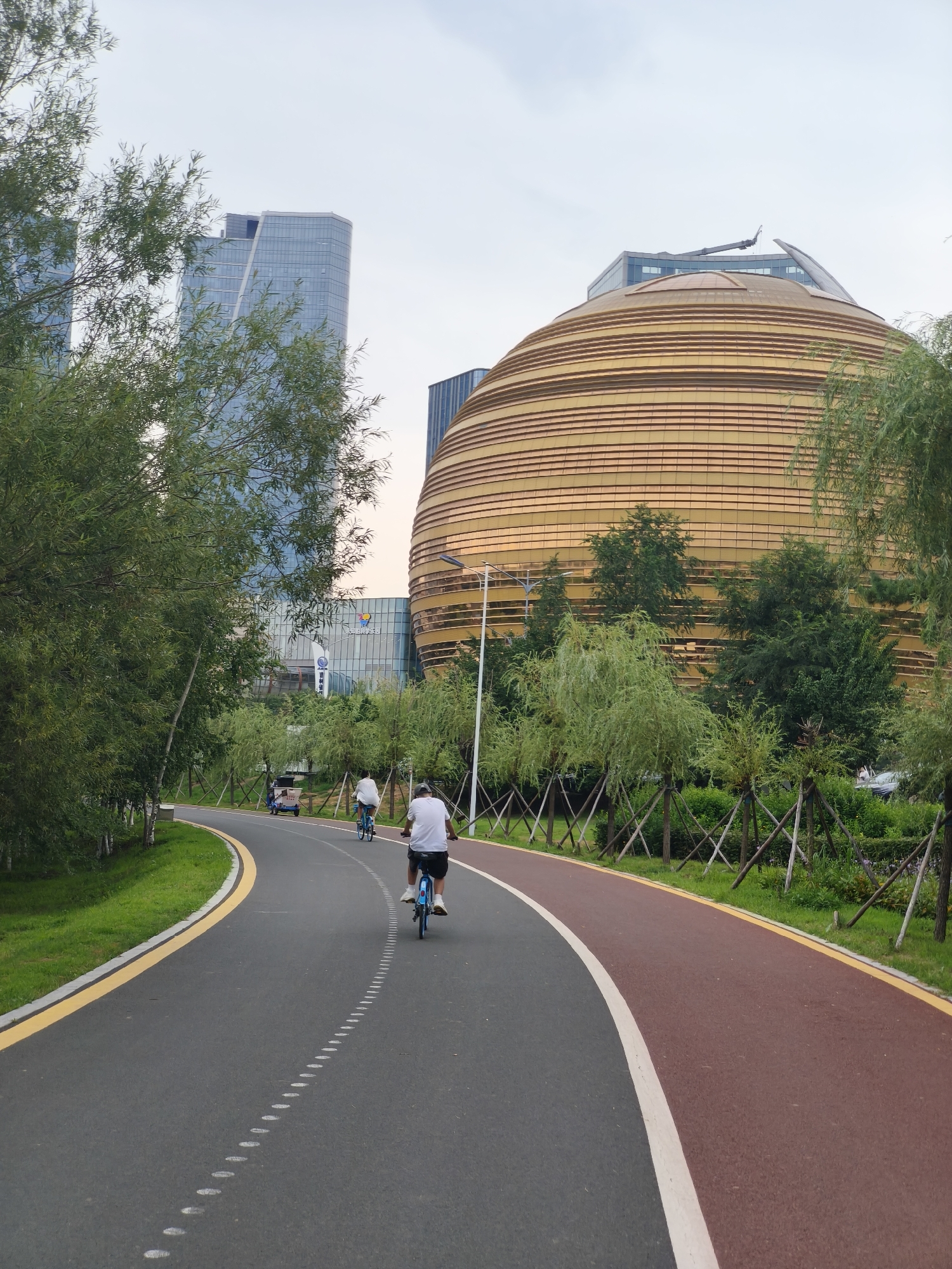 In Kuancheng District, China, a paved bike path curves through a park-like setting with trees and green spaces. The path is divided into a grey asphalt section for general use and a red section for bicycles. Two individuals are seen cycling on the grey path, moving away from the camera. In the background, a distinctive large, golden, spherical building with horizontal banding dominates the skyline, flanked by modern glass skyscrapers. The sky is overcast, suggesting a cloudy day.