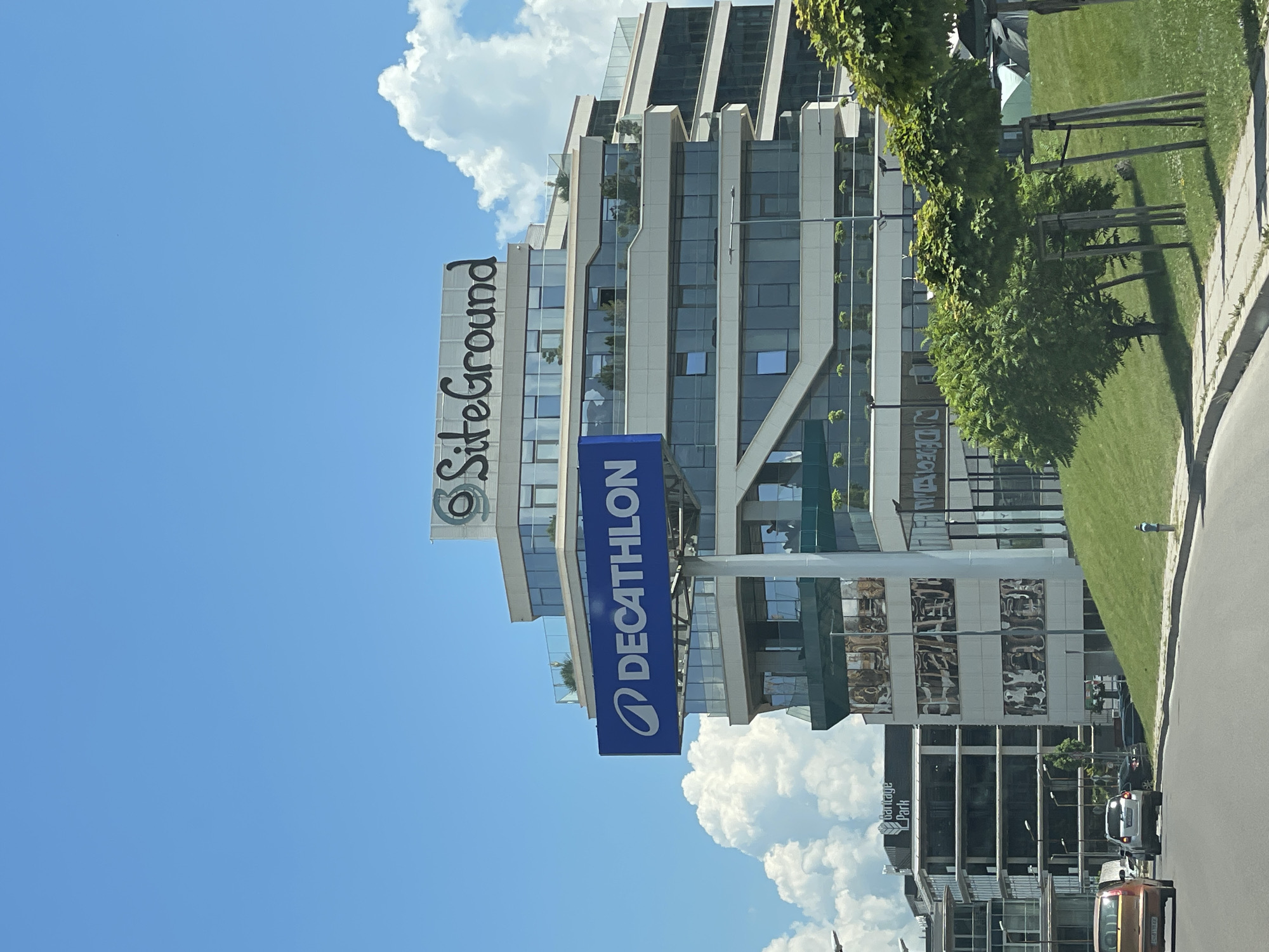 The image shows a modern office building with a large "DECATHLON" sign on a blue rectangular billboard mounted on a pole. Above the DECATHLON sign and on top of the building, a smaller sign reads "SiteGround" in a stylized font with a circular logo. The building features multiple levels with large glass windows and balconies with greenery. In the background, another building with "Garrige Park" signage is visible. The scene is set on a sunny day with a blue sky and scattered white clouds. A grassy slope with manicured trees is in the foreground, bordering a paved road where several cars are driving or parked. The location is identified as Sofia, Bulgaria.