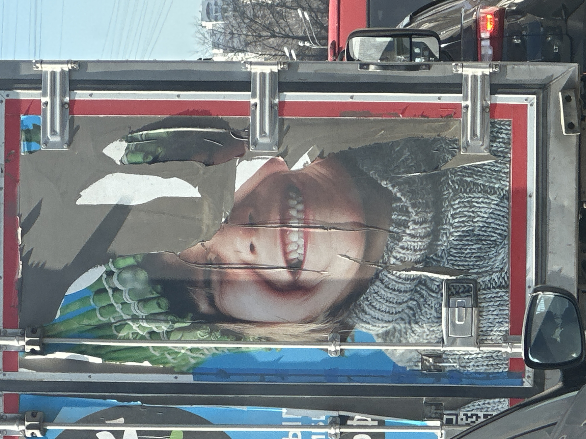 An advertisement poster is affixed to the back of a transport vehicle, possibly a truck or van. The poster depicts a woman's face with a pronounced smile and bright white teeth, held upside down, paired with images of asparagus. The poster is torn and worn. The vehicle is captured in transit with surroundings suggesting a city environment, specifically Moscow, Russia, due to the Cyrillic script visible on another poster below the first poster. Other vehicles and parts of the urban landscape are visible.
