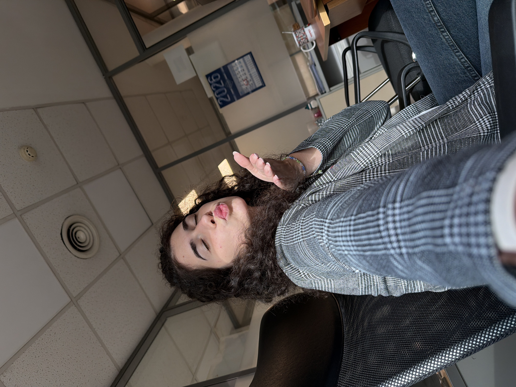 The image captures a woman with dark, curly hair sitting in an office chair and blowing a kiss towards the camera. She is wearing a grey plaid blazer and blue jeans. Her eyes are closed, and her lips are puckered. In the background, there are glass walls, a ceiling with ventilation and lighting fixtures, and a desk with a calendar showing the year 2026. The scene appears to be indoors, possibly in an office setting. The lighting suggests it is daytime, with sunlight casting shadows on the ceiling and walls.