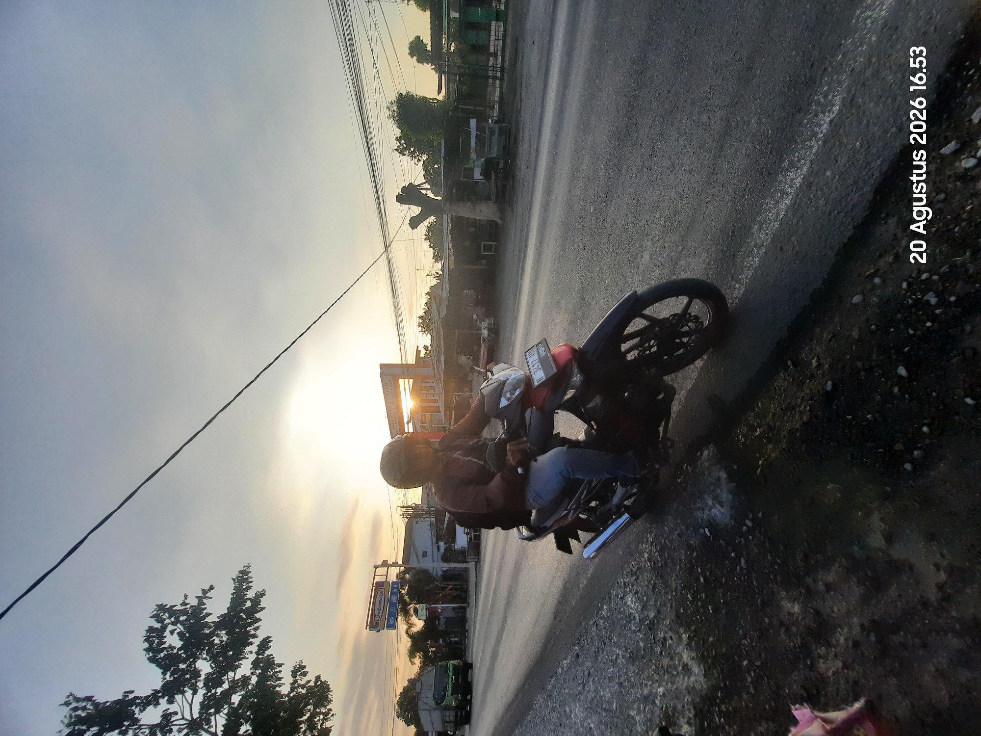 A person wearing a dark red long-sleeved shirt, blue jeans, and a black helmet rides a dark red and black motorcycle on a paved road in Sukokerto, Indonesia, at 4:53 PM on August 20, 2026. The person is carrying a black backpack. The motorcycle has a license plate that reads "3417 MH". A green truck is visible in the distance on the left side of the road, along with several buildings and stalls. The sun is setting in the background, casting a warm glow and long shadows. Overhead power lines crisscross the sky. The roadside is covered in gravel and some puddles of water.