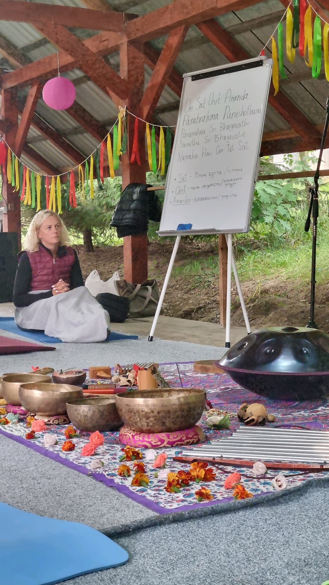 The image depicts an outdoor setting, likely a covered pavilion, in Izky, Ukraine, as suggested by the context. A woman with blonde hair is seated cross-legged on a blue yoga mat, wearing a dark long-sleeved shirt and a burgundy vest. She is looking upwards and to her left, with her hands clasped in her lap. In the foreground, a patterned rug is spread out, adorned with several brass singing bowls of varying sizes, small decorative objects, and scattered flower petals. To the right, a large, dark, metallic handpan rests on the rug. Behind the woman, a whiteboard on a tripod displays handwritten text, some of which appears to be in Sanskrit and Ukrainian. Colorful fabric streamers hang from the wooden structure above. The environment suggests a gathering for a meditation or sound healing session.