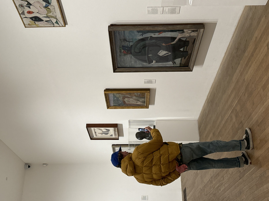 The image depicts an indoor gallery space, identified as the Tate Modern in London, United Kingdom, by the presence of Max Ernst's "The Elephant Celebes" (1921) on the right wall. A lone male individual, viewed from the rear, occupies the center-left. He wears a blue baseball cap, a mustard yellow quilted puffer jacket, light blue jeans cuffed at the ankles, and black low-top sneakers with white soles. He holds a dark-colored smartphone horizontally with both hands, aiming it towards a dark, abstract sculpted head encased within a clear rectangular plexiglass display, suggesting he is photographing or video recording the exhibit.
The gallery features white walls and a light brown hardwood floor. In addition to "The Elephant Celebes," several other framed artworks are visible: a smaller gold-framed painting to the left of "Celebes," a dark-framed abstract piece above the sculpture, and another abstract artwork in the upper right corner. Small information plaques are affixed to the wall next to certain art pieces. A white security camera is mounted in the upper left corner of the room.