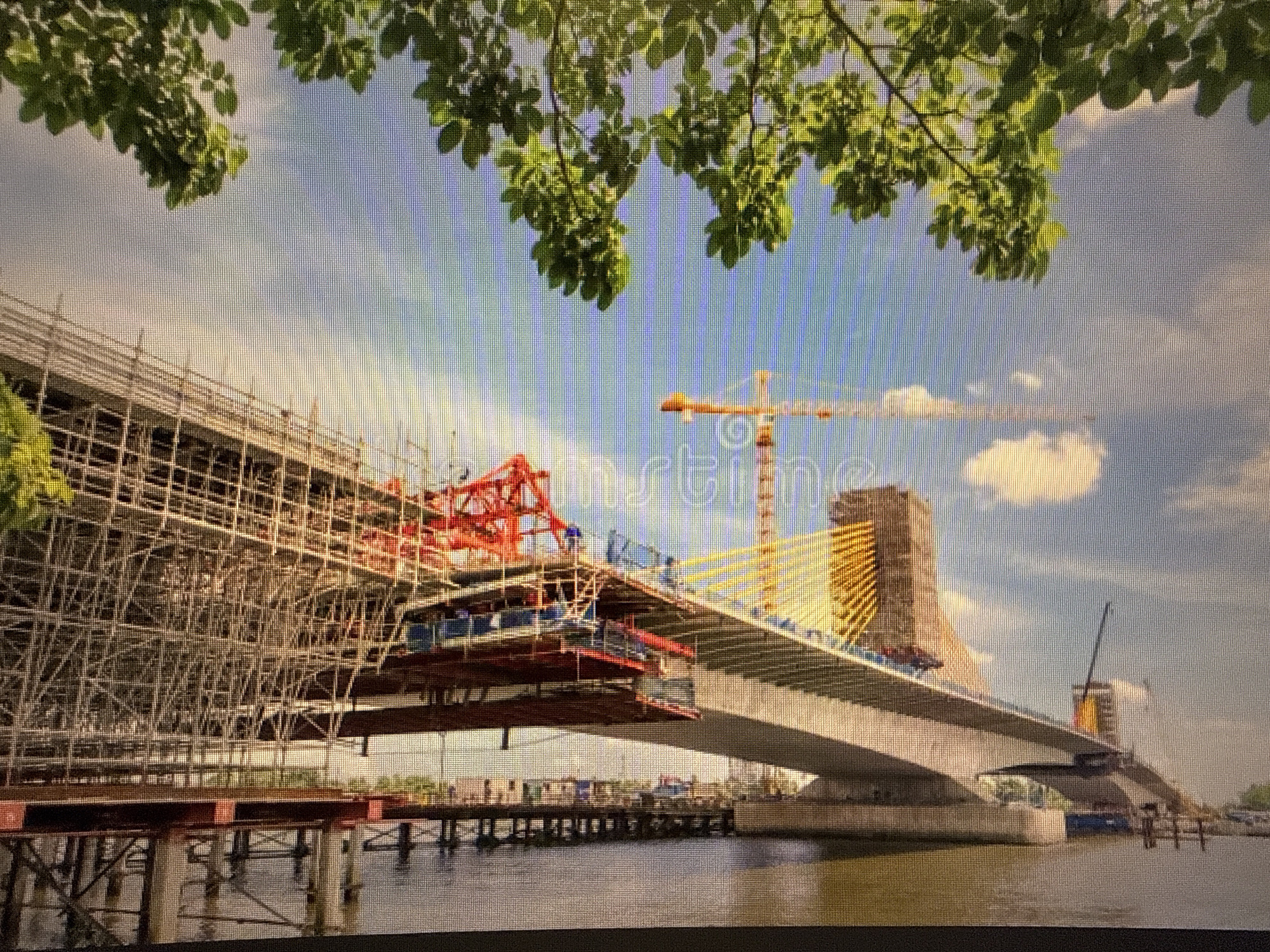 The image shows a large bridge under construction over a body of water, likely a river. Scaffolding surrounds a significant portion of the bridge, indicating ongoing work. A crane is visible in the background. There are temporary support structures in the water beneath the bridge. The sky is partly cloudy. The leaves of a tree are in the foreground, partially obscuring the view. The location is not Sofia, Bulgaria.