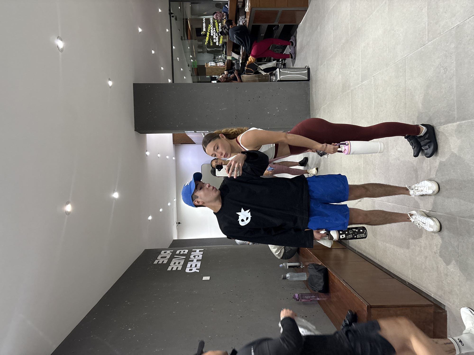 The image shows two people in what appears to be a gym or fitness studio. A person wearing a blue cap, black long-sleeve shirt with a white logo, and blue shorts is holding a phone, presumably taking a selfie. A woman is lying in a plank position behind them, wearing a white top and maroon leggings. She's using a white object, possibly a water bottle or a foam roller, for support. The floor is tiled, and a dark grey wall has white lettering, which seems to say "HIGHER VIBE". A wooden bench with various objects like water bottles and a gym bag is next to the wall. Other people are in the background. The location appears to be in San Andrés Cholula, Mexico.