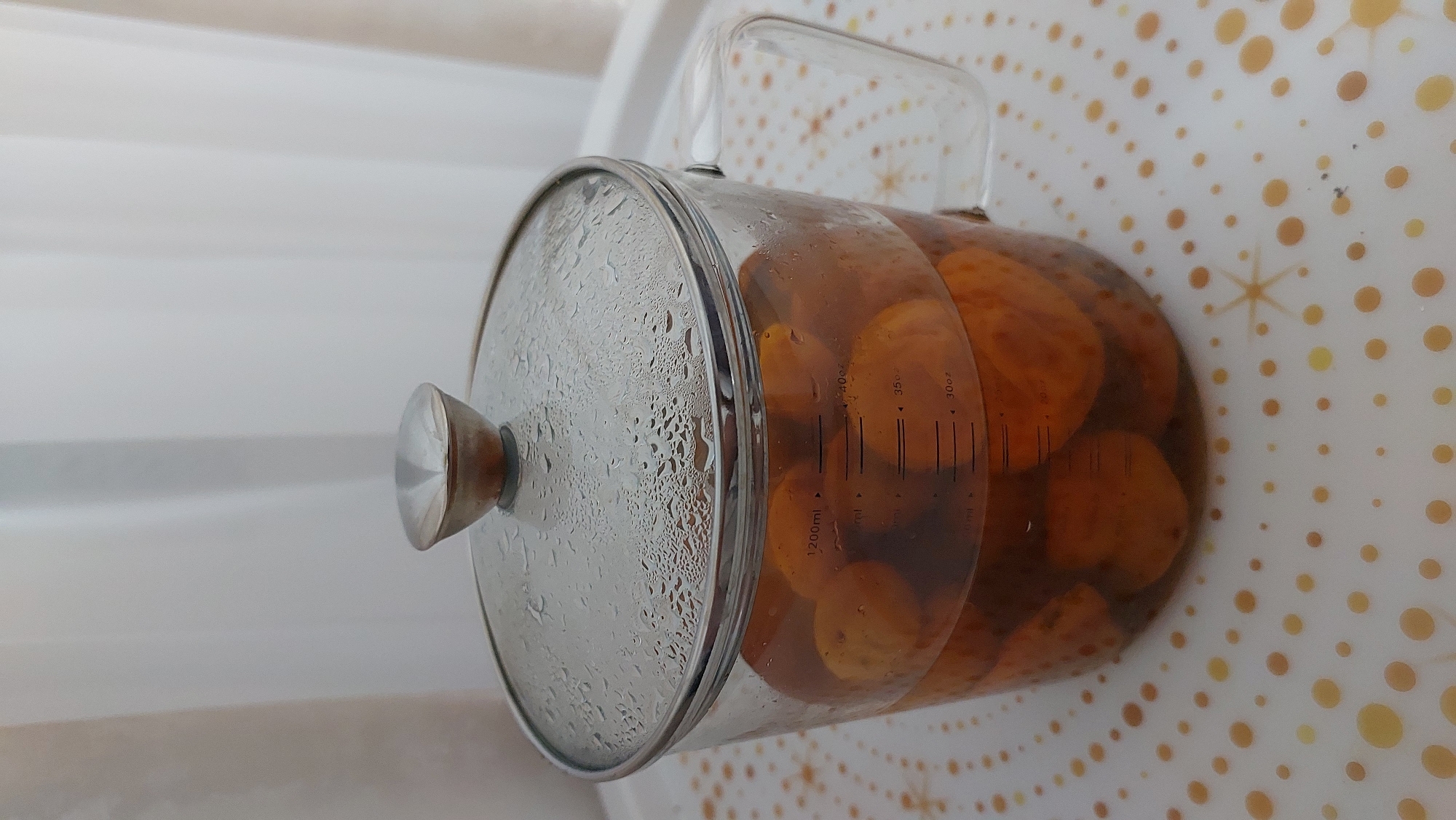 In Novocheboksarsk, Russia, a clear glass pot filled with amber-colored liquid and what appear to be whole apricots sits on a white surface adorned with a pattern of gold and brown circles and starbursts. The pot has a glass lid with a silver handle, and condensation droplets are visible on the underside of the lid. Measurement markings are visible on the side of the pot, indicating volumes up to 1200ml and in ounces. A glass handle is attached to the right side of the pot. The background consists of vertical white curtains. There are no people present in the image.