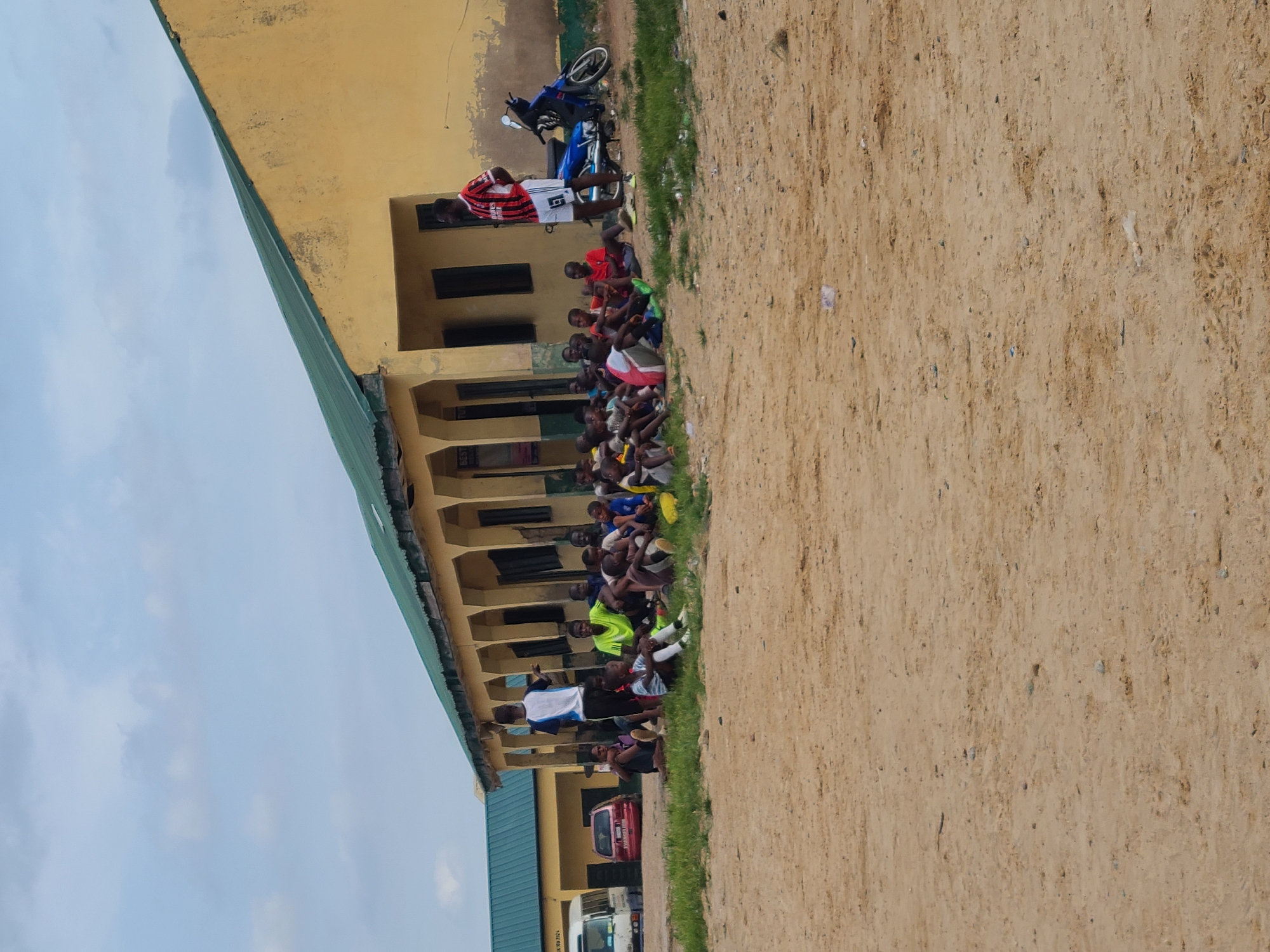 A group of young boys, some wearing soccer attire, are sitting on the ground in front of a yellow, single-story building with a green metal roof. A man in a red and white striped shirt and white shorts stands near a blue motorcycle parked to the right of the building. Several vehicles are visible in the background, including a white bus and a red car. The ground is sandy and there is patchy grass. The scene appears to be outdoors in a casual setting, possibly a school or community area in Masaka, Nigeria. The sky is cloudy.