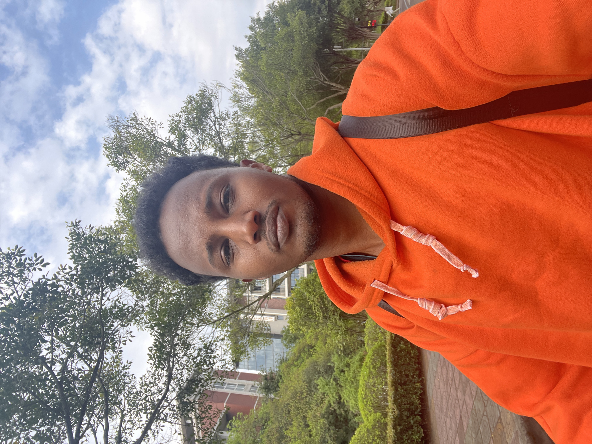 A medium shot depicts a man in an orange hoodie, with a dark brown strap over his shoulder, possibly for a bag. He is looking directly at the camera. The background includes trees, a brick-and-white building, and a cloudy sky. The man appears to be outdoors, likely in an urban area in Chongqing High-tech Zone, China.