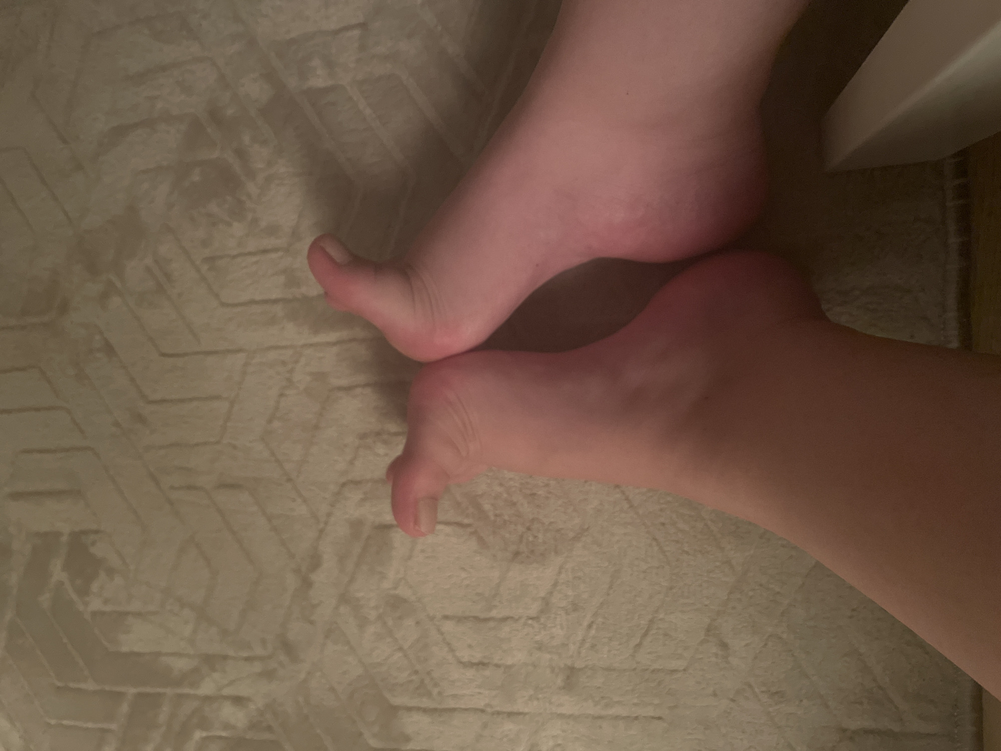 Two bare feet are shown from a top-down perspective, resting on a textured rug with a geometric pattern in a muted beige color. The feet are close together, with the toes of both feet pointing upwards and slightly to the left. The skin tone of the feet appears fair with a pinkish hue. The left foot is positioned slightly closer to the viewer and is curled upwards, with the toes clearly visible. The right foot is slightly behind and to the right of the left foot, and its sole is more prominently displayed. A sliver of a white object, possibly furniture, is visible in the bottom right corner of the image. The lighting appears to be dim, casting subtle shadows. The image is an indoor scene, and there is no indication of the specific location in Yunus Emre, Turkey, beyond it being an indoor setting.