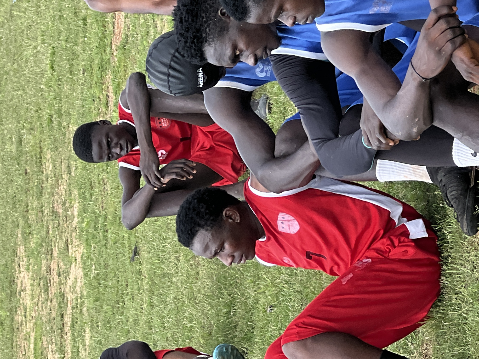 The image shows a group of young Black males sitting on a grassy field, appearing to be a sports team. The foreground features several individuals in athletic attire. One person on the left wears a red jersey with white trim and the number "7" prominently displayed. Another individual in the center wears a black swim cap with "ARENA" written on it and a blue and white athletic top. To the right, more individuals in blue and white athletic wear are visible, with one wearing black sleeves. The grass is green with some patches of dried brown. In the background, another individual in a red jersey with white trim is seated, looking forward. The lighting suggests an outdoor, daytime setting. The image does not provide specific visual cues to confirm its location within Jos, Nigeria, but the overall context suggests a sports training or game scenario.