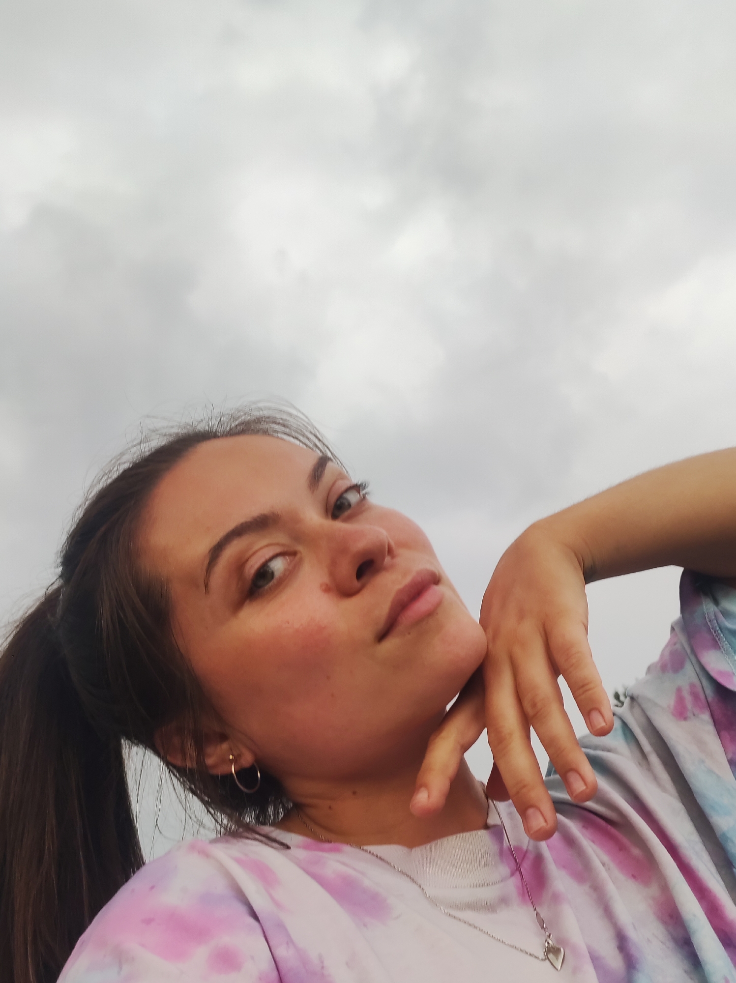A young woman is captured in a close-up, low-angle selfie against a backdrop of a cloudy sky. She has dark hair pulled into a ponytail, and she is wearing a tie-dye t-shirt with pink, blue, and white patterns. A silver necklace with a heart-shaped pendant is visible around her neck. Her left hand is resting on her chin, and she is looking directly at the camera with a relaxed expression. The lighting suggests it might be overcast or late afternoon. The image does not provide any specific clues to pinpoint its location within the city of Фоки, Russia.