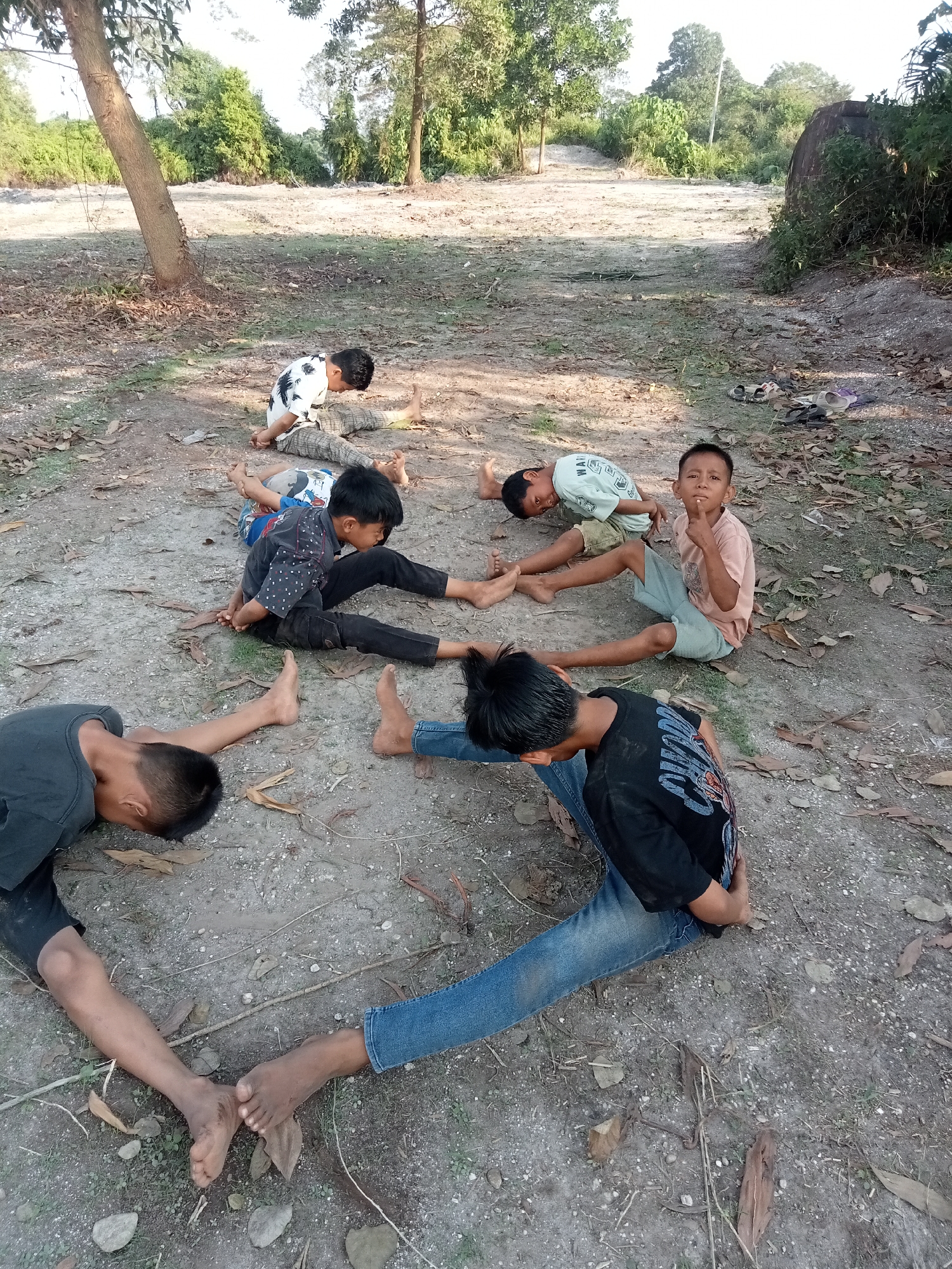 The image depicts a group of seven boys, appearing to be of school age, arranged in a circle on a dirt path. They are all lying down or sitting in various yoga-like poses. The environment is outdoors, with dry leaves and dirt covering the ground. Trees and foliage are visible in the background, suggesting a rural or natural setting. One boy in the center is sitting upright and looking at the camera, with his index finger raised to his lips in a gesture of silence. The other boys are mostly facing down or away from the camera, engaged in their respective poses. The overall scene appears to be a recreational or educational activity taking place in Teluk Kenidai, Indonesia.