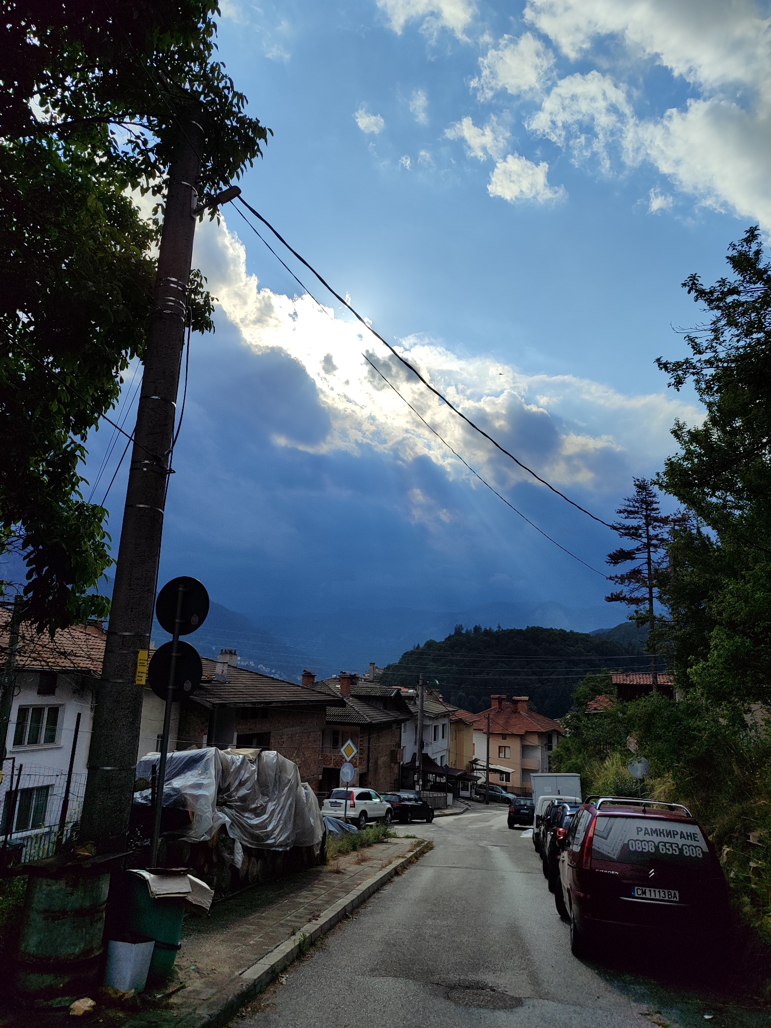 The image depicts a residential street in Smolyan, Bulgaria, on a cloudy day with sunlight breaking through the clouds. The street is paved and slopes slightly upwards. Several parked cars are visible along the right side of the street, including a dark red Citroën with Bulgarian license plates and text in Cyrillic script on its rear window. Houses with red-tiled roofs line both sides of the street. A tall utility pole with wires runs along the left side of the image, partially obscuring the view of the sky. Trees and lush greenery are present, particularly on the right side of the street and in the background, where hills covered in vegetation are visible. The sky is a mix of blue and dark clouds, with rays of sunlight casting dramatic shadows. There are no people visible in the image. The scene suggests a quiet, everyday moment in a town or village setting.