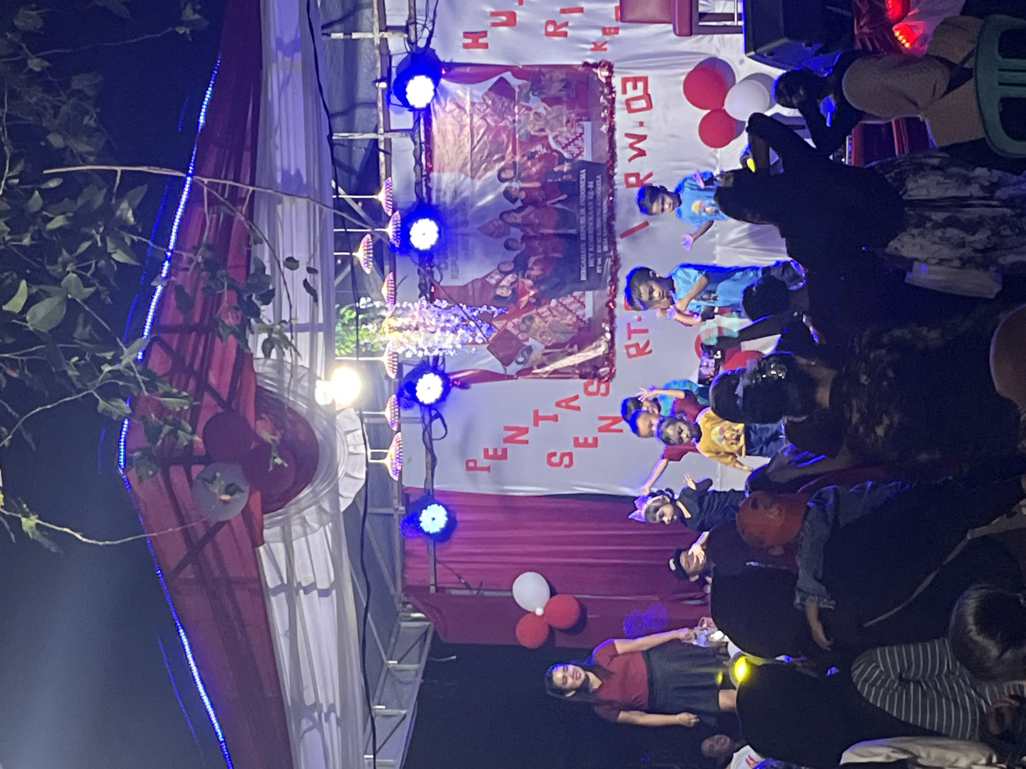 This image depicts an outdoor stage event in Tajurhalang, Indonesia, likely a community gathering or performance. A group of children are on stage, some dressed in colorful clothing, appearing to participate in a performance. One child in a yellow shirt is holding a microphone, and another in a blue shirt is also holding a microphone. Behind them, a banner displays "PENTAS SENI" and the text "REPUBLIK INDONESIA HUT KEMERDEKAAN KE-77". Red and white balloons are visible on stage and to the side. A woman in a red shirt and black skirt is standing on the left side of the stage, observing the performance. The stage is decorated with red drapes and illuminated by blue stage lights. The event appears to be taking place at night, with foliage hanging from above the stage. A crowd of people are gathered in front of the stage, some observing the event, others interacting with each other. Some individuals in the foreground are holding up cell phones, likely to record the performance. The overall atmosphere seems festive and community-oriented.
