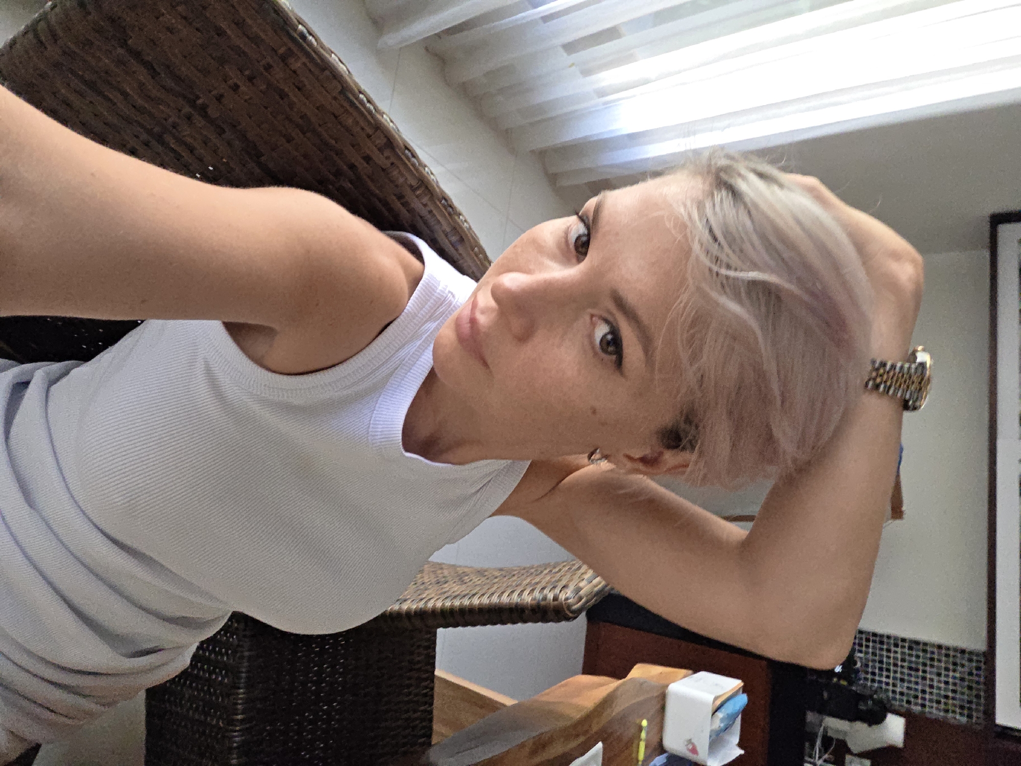 The media file is a selfie of a woman with short, light-colored hair and fair skin. She is wearing a white tank top. She is leaning on a wicker chair and looking towards the camera, with her head tilted. Her left arm is resting on the back of the chair, and her right arm is bent, with her hand near her head. She is wearing a watch on her right wrist. The background shows a ceiling with lights, a wall with patterned tiles, and a framed picture on the wall. The lighting suggests it is daytime. The scene appears to be indoors, possibly in a tropical location given the mention of North Kuta, Indonesia. The woman's expression is neutral. There is no visible text in the image.