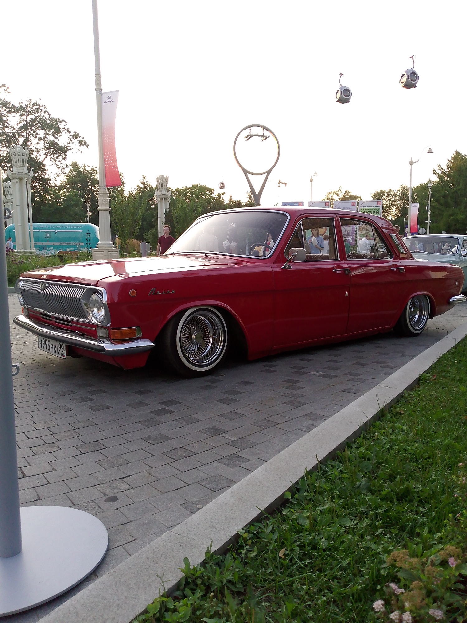The image captures a vintage red Volga GAZ-24 sedan parked on a cobblestone path in a city park in Moscow, Russia. The car, lowered and customized with wire-spoke wheels, is the primary subject. In the background, a man in a dark t-shirt stands near a turquoise bus. Further back, trees and architectural elements, including decorative lampposts and banners, suggest an urban park setting. In the sky, two spherical objects, possibly decorative lights or part of a public art installation, are visible. The ground is paved with cobblestones and bordered by a grassy area. The overall lighting suggests it is late afternoon or early evening.