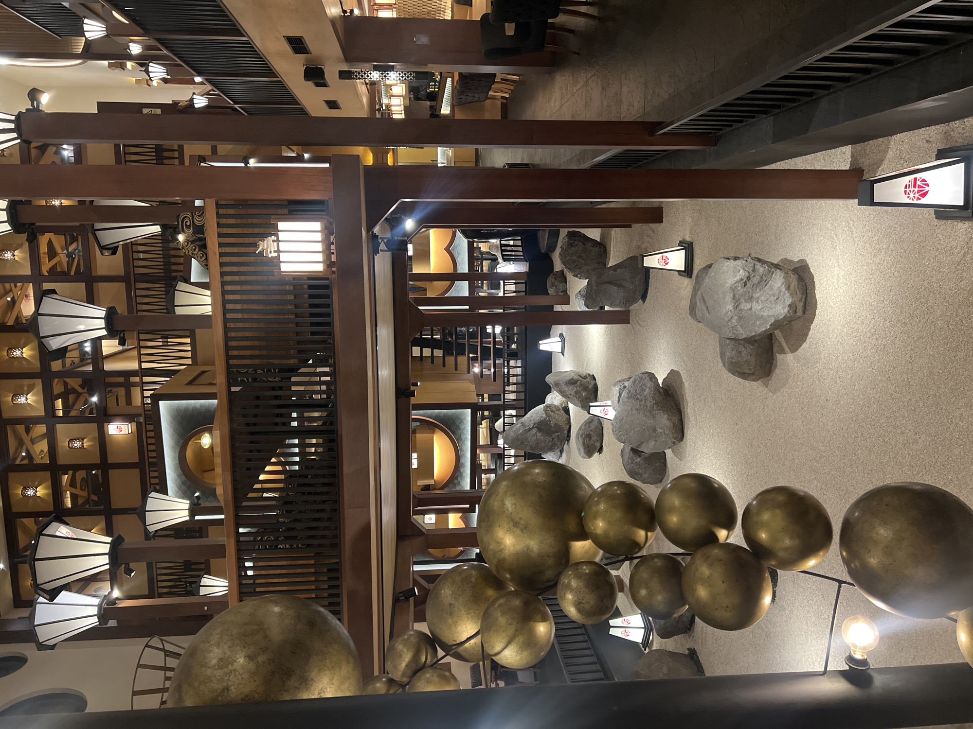 The image shows the interior of a building in North Kuta, Indonesia. The interior design features a blend of modern and traditional Indonesian elements. The building has multiple levels, with wooden beams and latticework forming the architectural structure. There are several decorative elements, including large golden orbs hanging from the ceiling, and large grey rocks as part of the interior design. There are lights with white shades, and lanterns with red and white signs. In the background, there are visible tables and chairs, indicating a dining or seating area. A staircase is visible leading to upper floors. The environment appears to be well-lit and spacious.