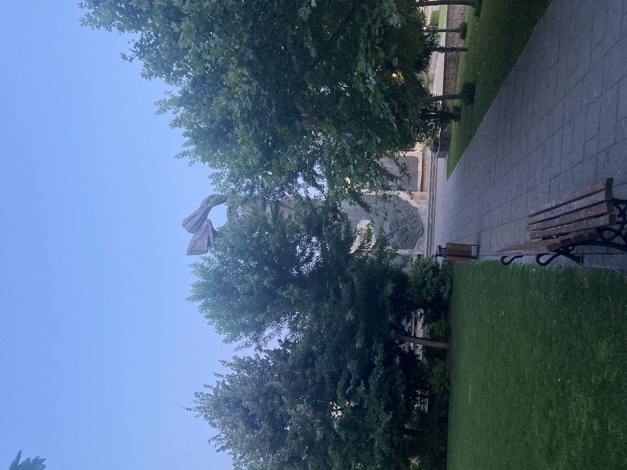 The image depicts a wide, paved walkway leading towards a large, abstract monument in a park-like setting. The sky is a clear, pale blue, indicating it may be early morning or late evening. Lush green trees flank both sides of the walkway, with their leaves casting dappled shadows. A wooden bench is positioned in the foreground on the left side of the path, and a trash receptacle is visible further up on the right. The monument itself is a towering structure with angular, stone-like elements, and intricate carvings or relief work visible at its base. The overall impression is one of a public space, likely a memorial or landmark, within a city. This scene is located in the city of Iași, Romania, specifically at the Monumentul Eroilor Neamului (Monument to the Heroes of the Nation), also known as the Cenotaph.