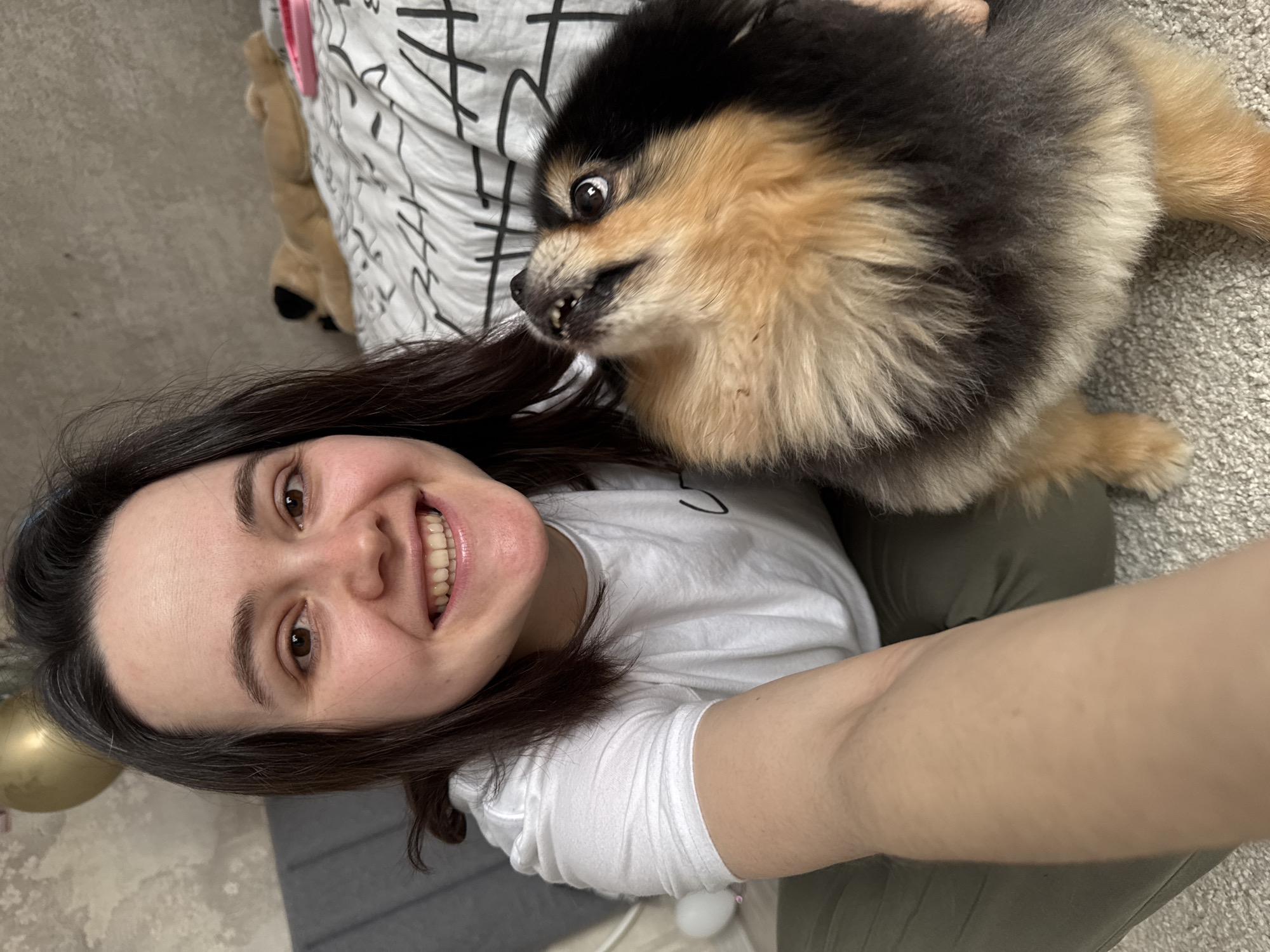 A smiling woman with brown hair and brown eyes is taking a selfie with a Pomeranian dog. The woman is wearing a white t-shirt and olive green pants. The dog, which appears agitated, has black and tan fur and is biting at the woman's hair. They are positioned on a carpeted floor, with a bed or other soft surface covered in a white patterned sheet behind them. In the bottom left of the frame is part of a gold-colored balloon. It's not possible to identify the location as Izhevsk, Russia from the given image.