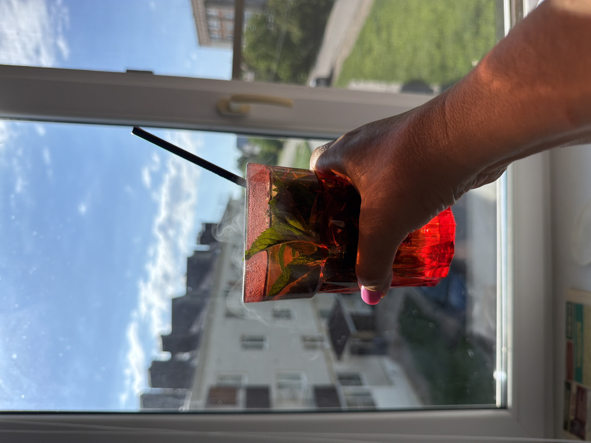 A hand holds a red glass filled with a dark liquid and green leaves near a window. A black straw is visible in the glass. A wisp of vapor rises from the drink. Outside the window, buildings are visible, along with a blue sky with clouds. A city block is seen beyond the window. The location is Leningradskaya, Russia.