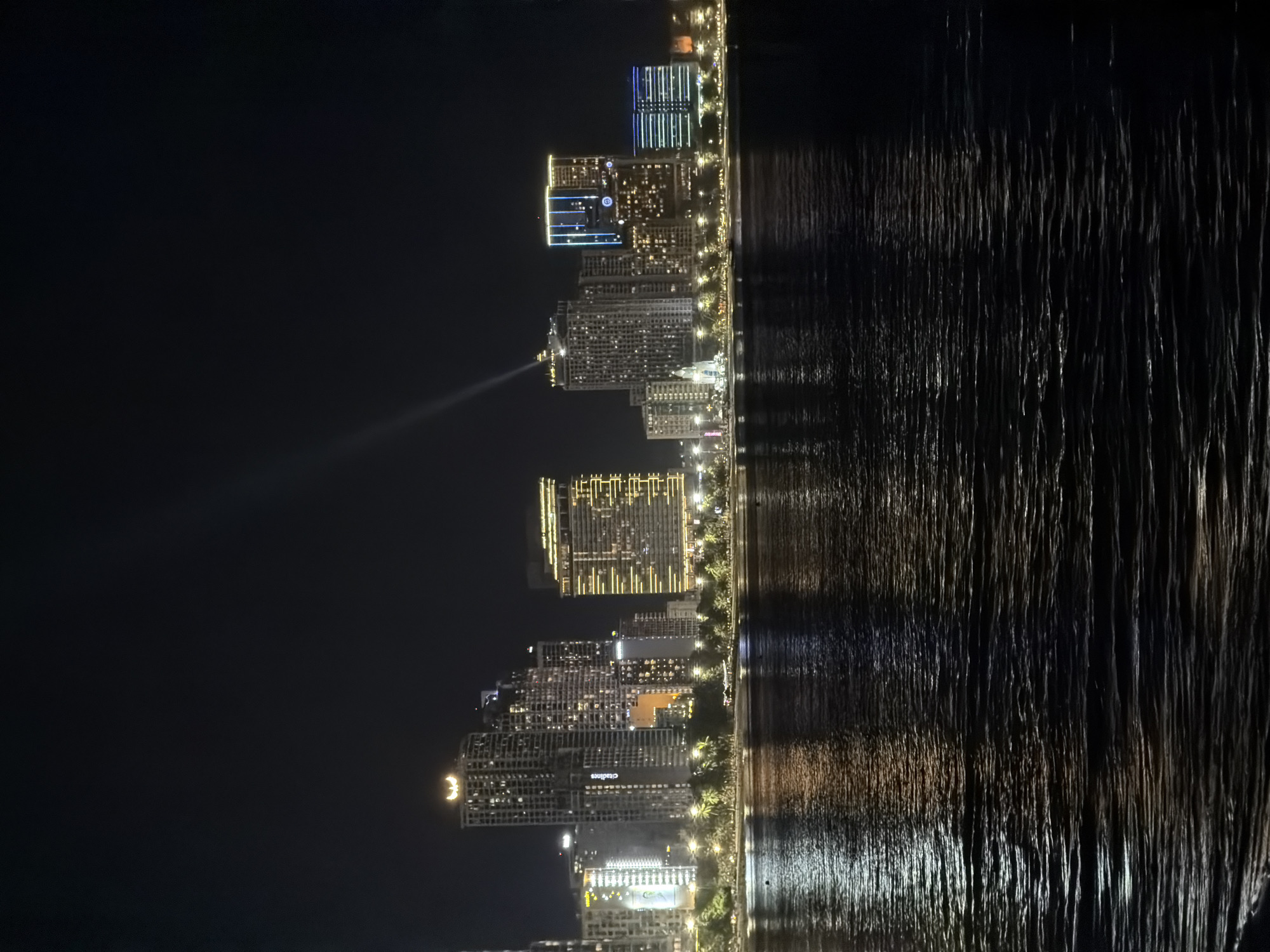 The image captures a nighttime view of a coastal city skyline in Nha Trang Ward, Vietnam, dominated by numerous illuminated high-rise buildings and their reflections on the dark water in the foreground. The buildings display a variety of architectural styles and lighting schemes, ranging from uniformly lit windows to accent lighting that highlights architectural features. One prominent building on the left has a distinctive crescent moon logo illuminated at its peak. A powerful beam of light is emanating from the top of another building towards the night sky. Along the waterfront, a line of trees and smaller structures are visible, also lit from within or by streetlights, contributing to the overall ambiance of the urban setting. The water surface is textured with ripples, creating a shimmering reflection of the city lights. No people are visible in the image.