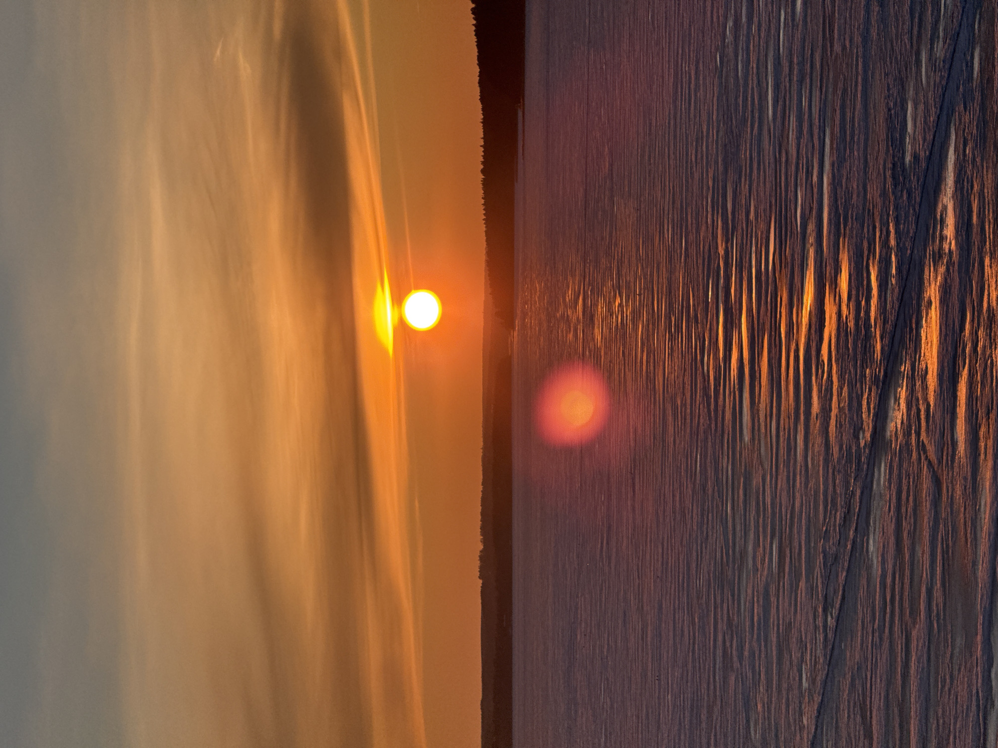 A vertical photograph depicts a sunset over what appears to be a body of water, likely taken in Votkinsk, Russia. The sun, a bright orange orb, is positioned in the upper left quadrant of the frame, casting a diffuse glow onto the sky to its left. To the right of the sunset there is a band of silhouetted trees that separates the water from the sky. The water occupies the right half of the image, with the setting sun reflecting off the ripples and currents on the surface. The reflection gives an elongated, diffused light. The water's texture is marked by linear patterns that suggest movement and surface disruption. The sky presents a soft gradient of colors, transitioning from light to dark.
