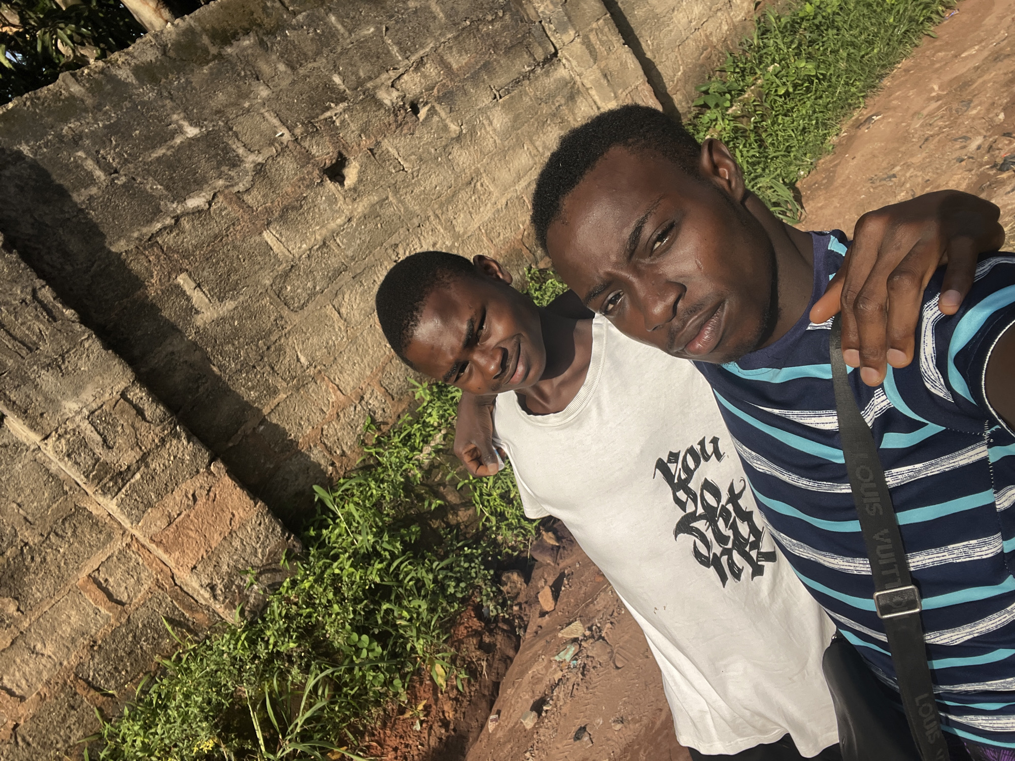 Two young men are captured in a selfie-style photograph, their faces turned towards the camera. The man on the right is closer to the lens, his head tilted slightly and his eyes looking directly at the viewer. He is wearing a dark blue and white striped t-shirt and a black strap from a bag crosses his chest. The man on the left is partially behind him, smiling and looking slightly off-camera. He is wearing a white t-shirt with a graphic print.
The background suggests an outdoor setting in Nkpor, Nigeria. A rough, textured brick wall forms the backdrop on the left, with patches of green vegetation growing around its base. To the right, the ground is a mix of dry, reddish-brown earth and more green plants. The lighting indicates it is daytime, with strong sunlight casting shadows. The overall mood appears casual and friendly. The strap of the bag has visible text which reads "LOUIS VUITTON" repeated.