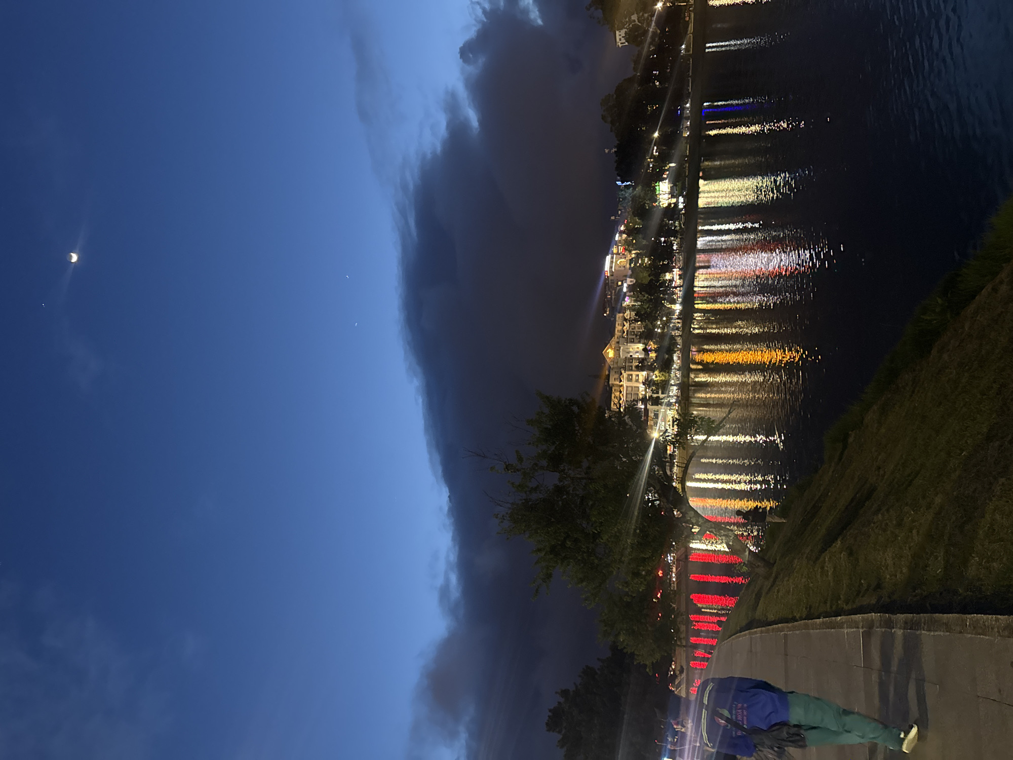 This nighttime image depicts a scene in Da Lat, Vietnam. The sky transitions from a deep blue with a crescent moon to a dark, stormy cloud formation. Adjacent to the cloud mass, lights from buildings illuminate the city's waterfront, reflecting in the water. Along the edge of the waterway, there's a promenade with lights, trees, and a grassy bank. A person dressed in blue and green clothing lies on the pavement.