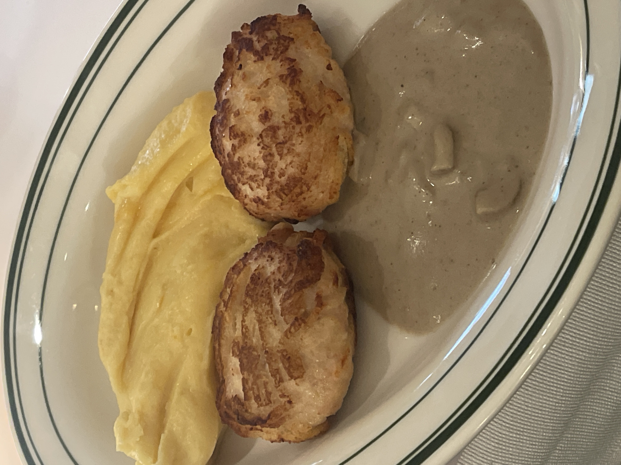 A close-up shot shows a white plate with green stripes, filled with food. On the left side of the plate is a portion of creamy, bright yellow mashed potatoes. To the right of the mashed potatoes are two small, round, golden-brown patties, appearing to be fried or pan-seared. Their surfaces show a slightly uneven texture and some darker brown crispy bits. The right side of the plate contains a pool of light brown, smooth sauce, possibly a mushroom or gravy sauce, with a few pale pieces of what appear to be mushrooms visible within it. The food is presented on a white surface, likely a table, with a textured gray cloth visible at the bottom right of the frame. The lighting suggests it is daytime, possibly indoors, and the overall presentation is of a prepared meal. There is no visible text in the image.
