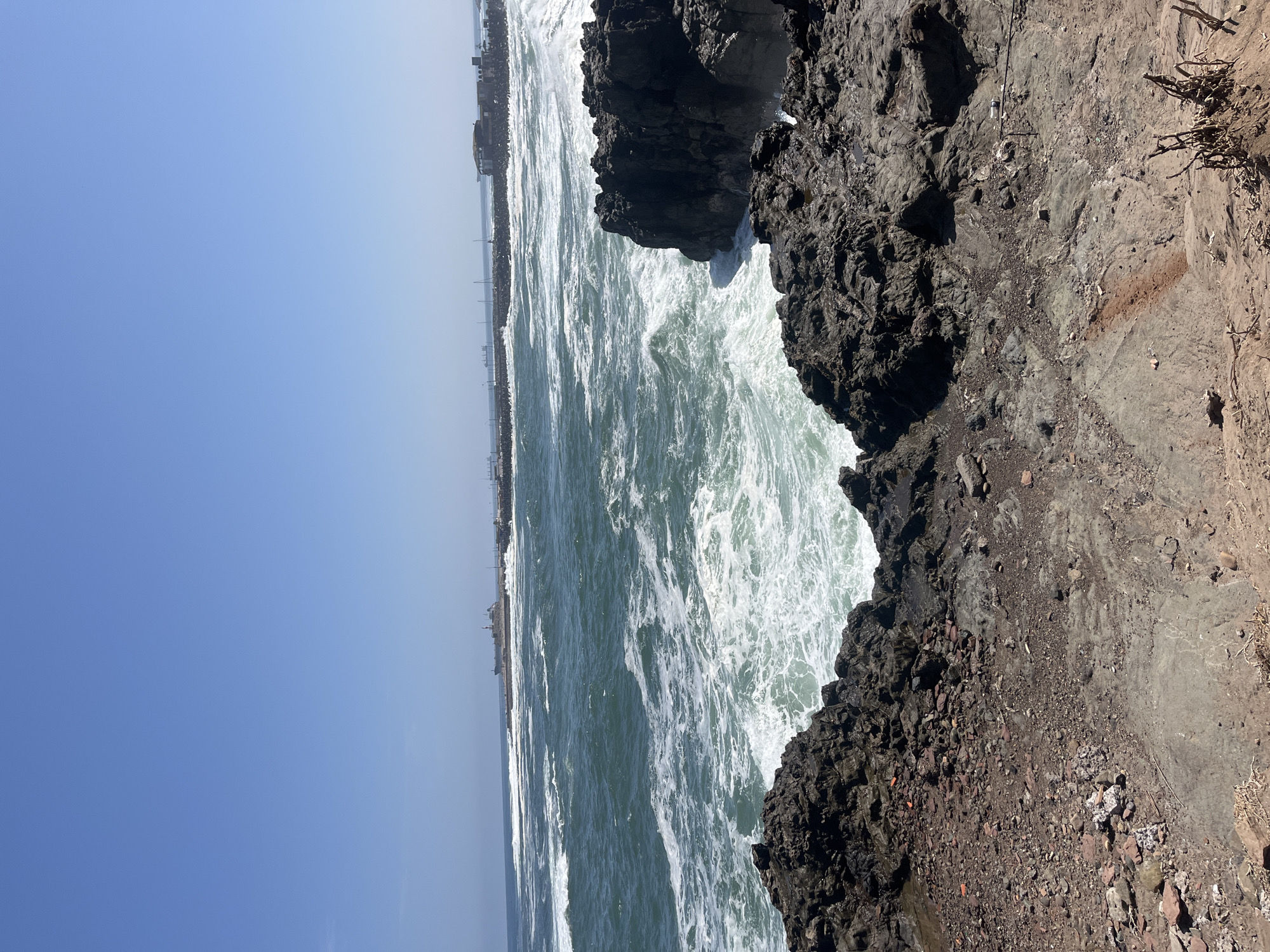 The image depicts a coastal scene in Arica, Chile, featuring a rocky shoreline and the Pacific Ocean. The ocean, a vibrant mix of green and white, is characterized by waves crashing against the rocks. The rocky terrain is rough and dark in color. In the background, along the horizon, is a cityscape with buildings and structures. The sky above is a clear, bright blue. The vantage point is close to the edge of the rocks, creating a sense of proximity to the ocean's power. There are no people visible in the frame.