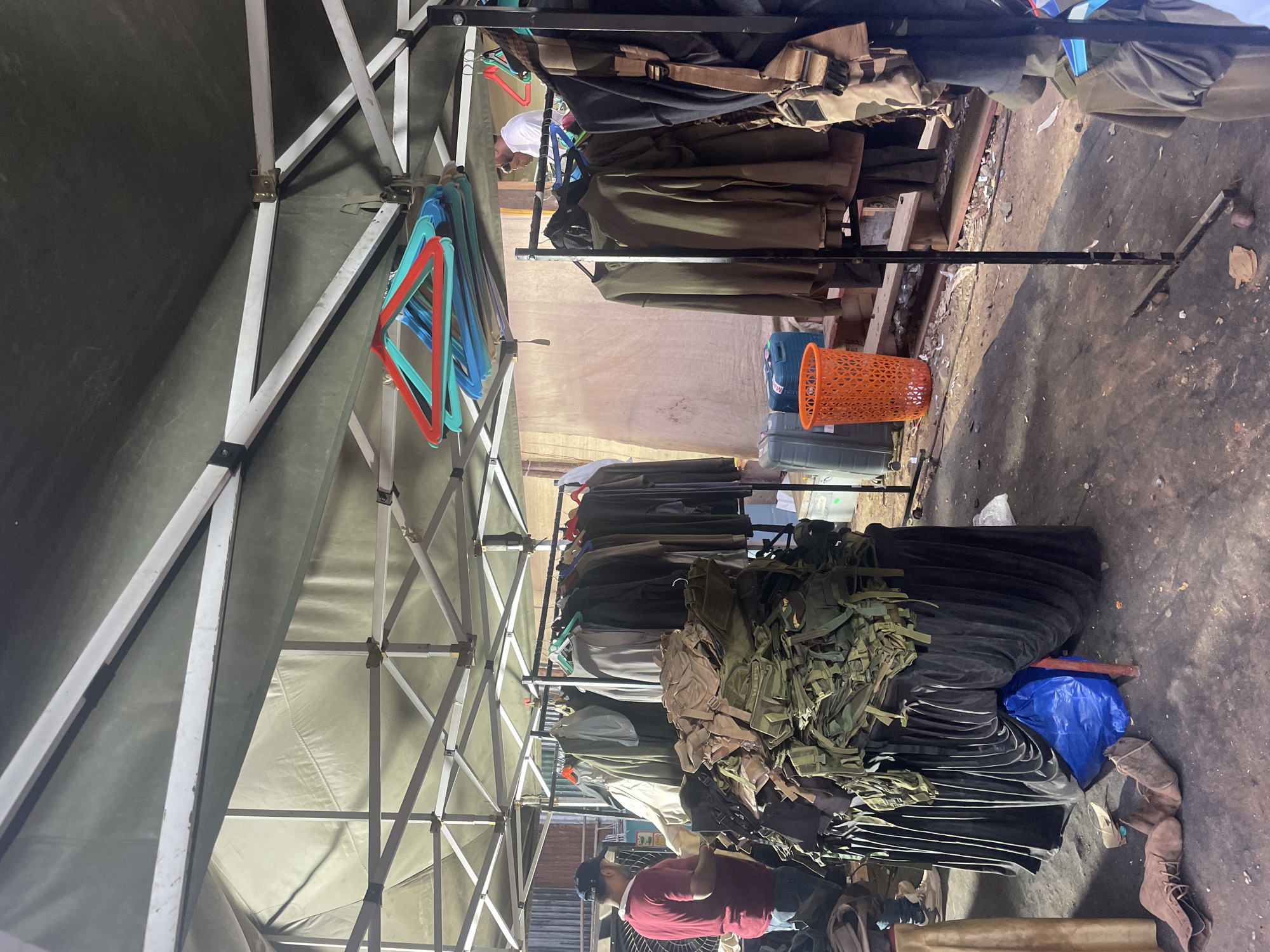 The image depicts an outdoor retail setup, possibly a market stall in Mira-Bhayander, India. Garments, appearing to be military-style clothing and accessories, hang on metal clothing racks under a white and gray pop-up tent. Below the hanging clothes, piles of folded dark-colored fabrics and olive drab gear are visible. A blue plastic bag sits on the ground, along with a pair of brown boots. A person wearing a red shirt and a baseball cap stands partially obscured near the bottom of the frame. An orange plastic basket is partially visible. The ground is a dark, rough surface. The light is diffused, suggesting an overcast day or a shaded area.