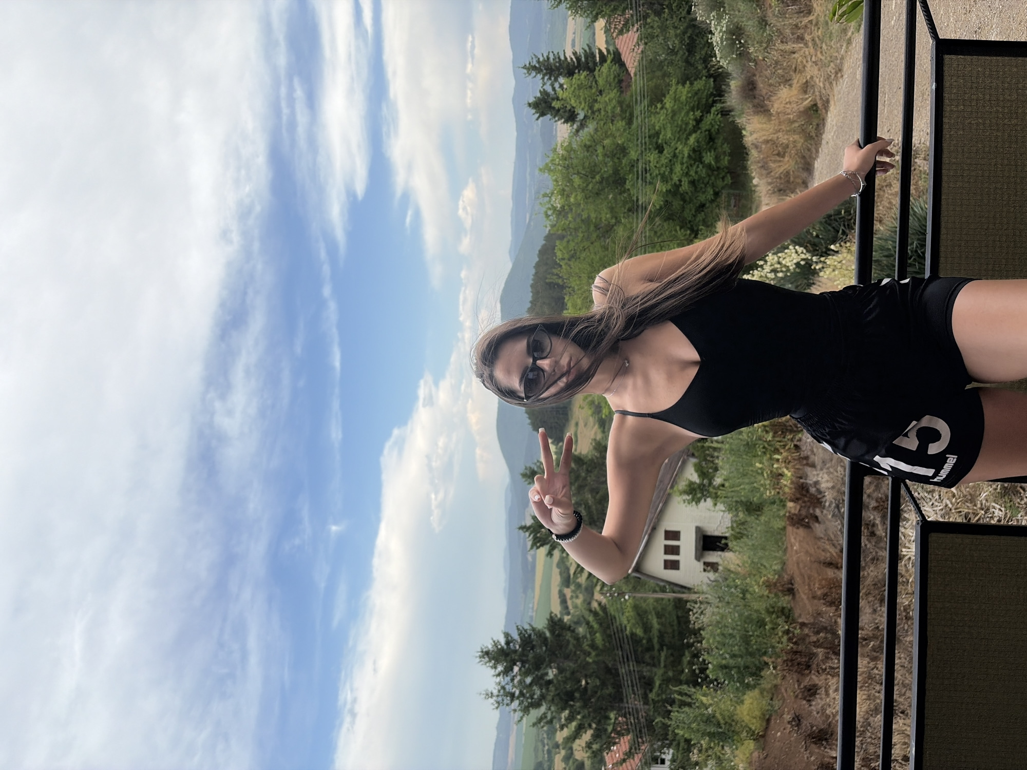 A young woman with long brown hair and sunglasses is posing for a photo on a balcony. She is wearing a black tank top and black shorts with "hummel 13" visible on the right leg. She is holding up two fingers in a peace sign with her right hand, and her left arm is resting on the balcony railing. The background shows a scenic view of rolling hills covered in trees and a small white house. The sky is partly cloudy with blue skies. The time of day appears to be daytime. The location is Vladislavtsi, Bulgaria. The woman appears happy and relaxed.